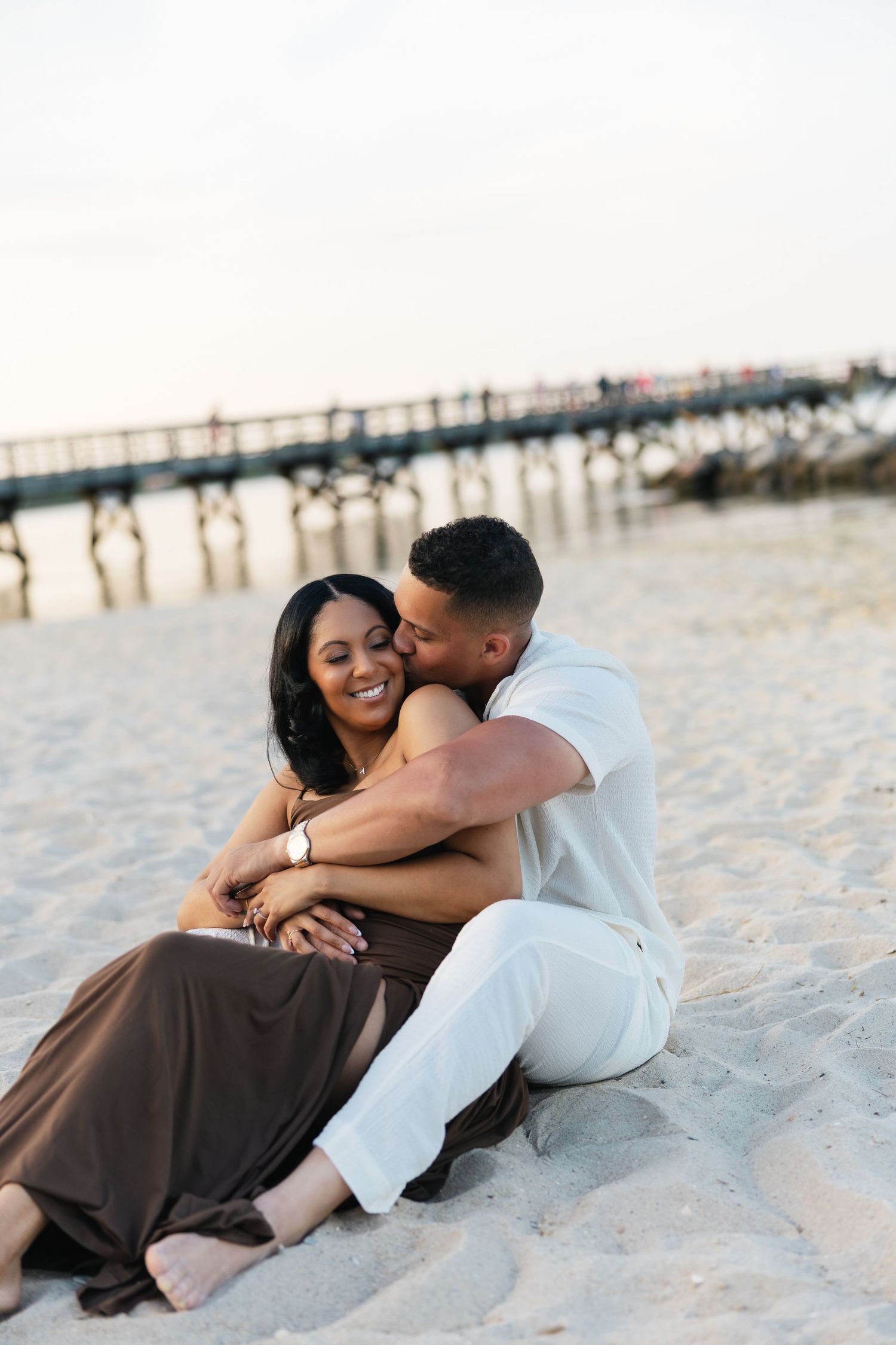 Couple sitting on beach sand, man kissing woman's cheek, pier in background at sunset.