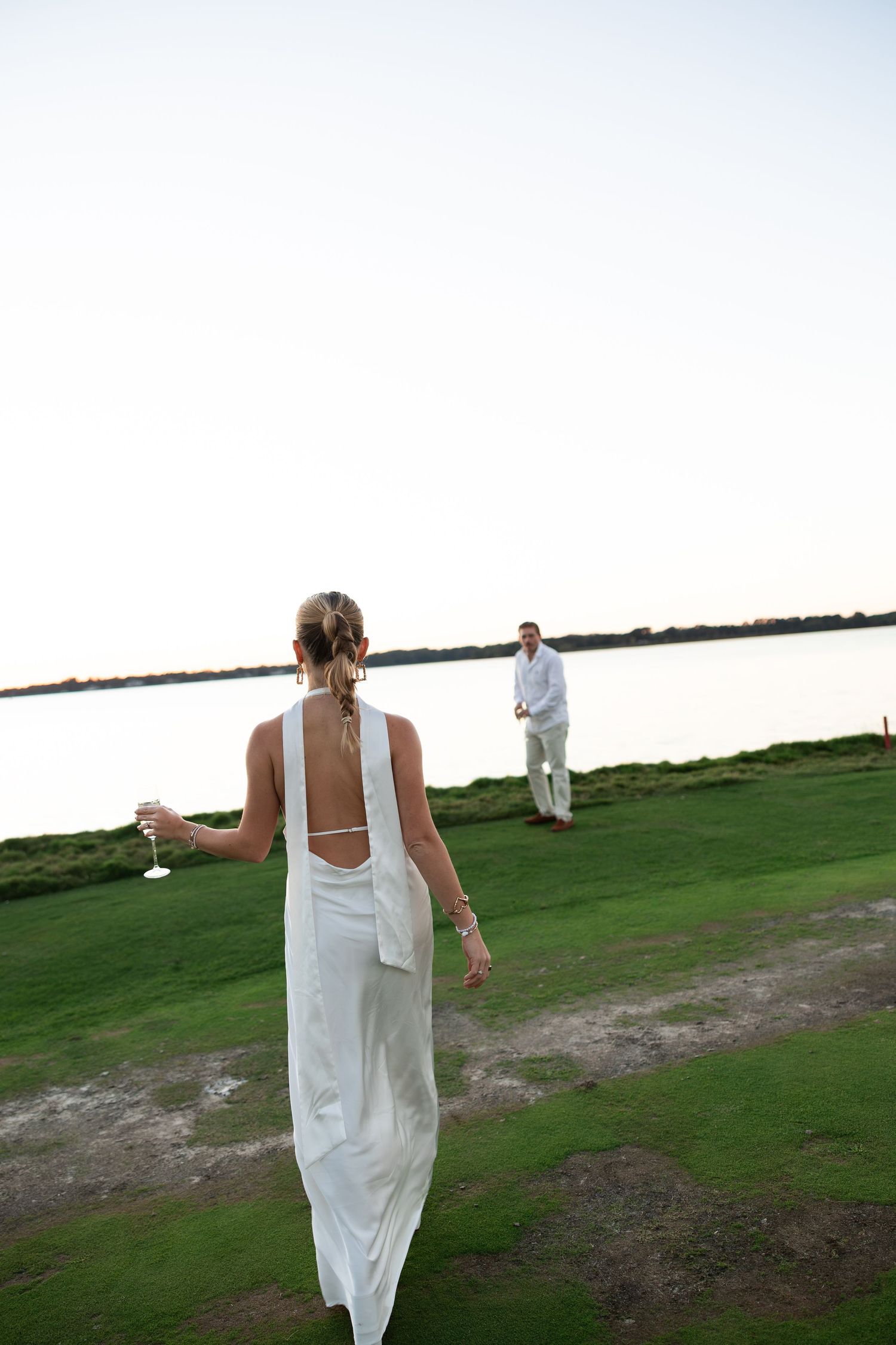 Bride in white backless gown walks toward groom on green lawn by waterfront at dusk.