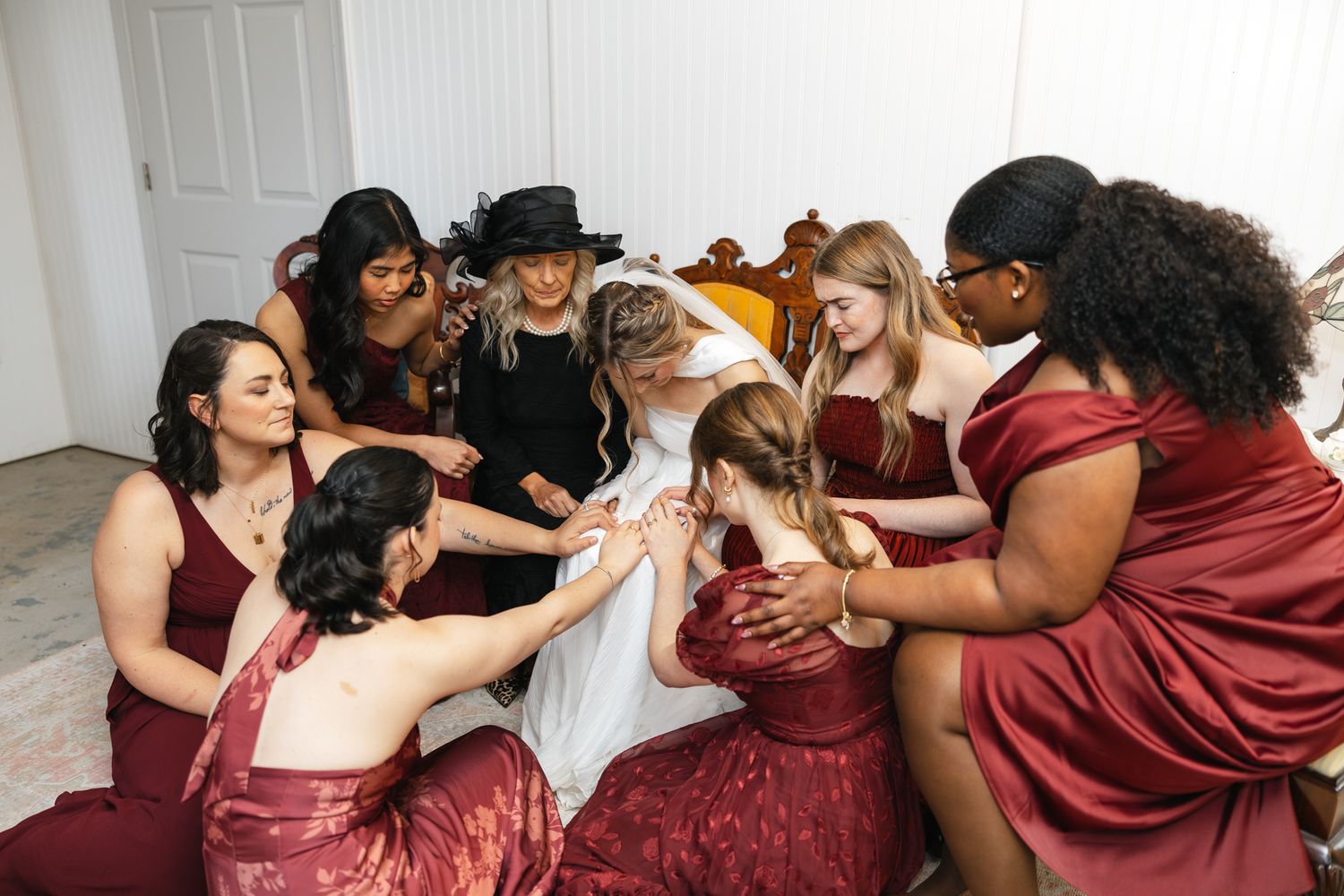 Bridesmaids in burgundy dresses gather around bride in white gown for group prayer before wedding.