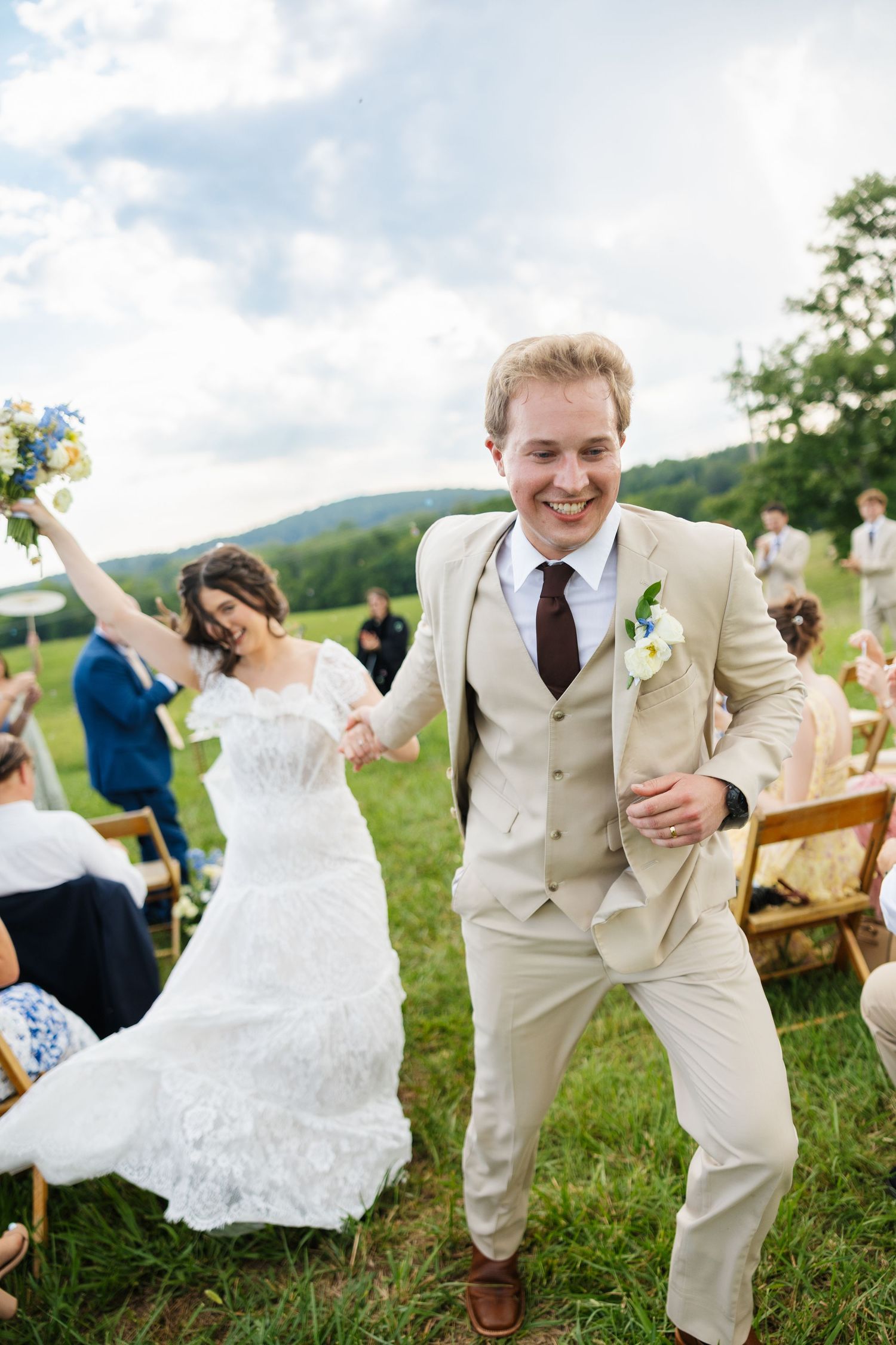 Joyful groom in tan suit leads bride in white lace dress down outdoor wedding aisle on green lawn.