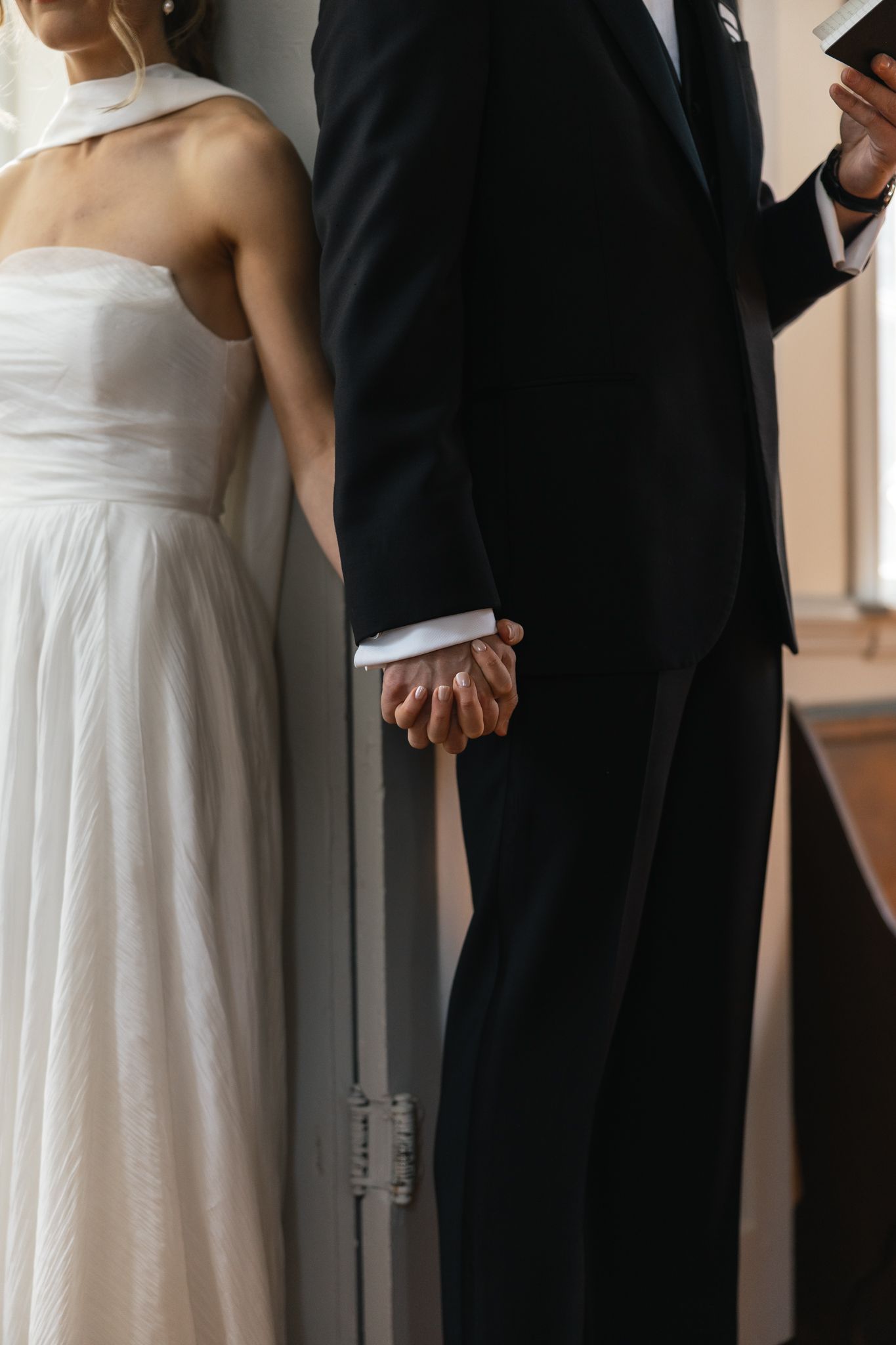 Bride in white halter gown and groom in black suit hold hands tenderly during intimate wedding moment.