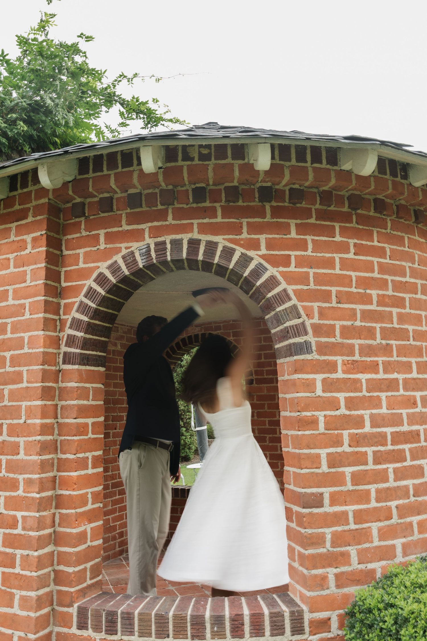 Bride in white dress and groom in dark jacket pose under a brick archway gazebo in an outdoor garden setting.