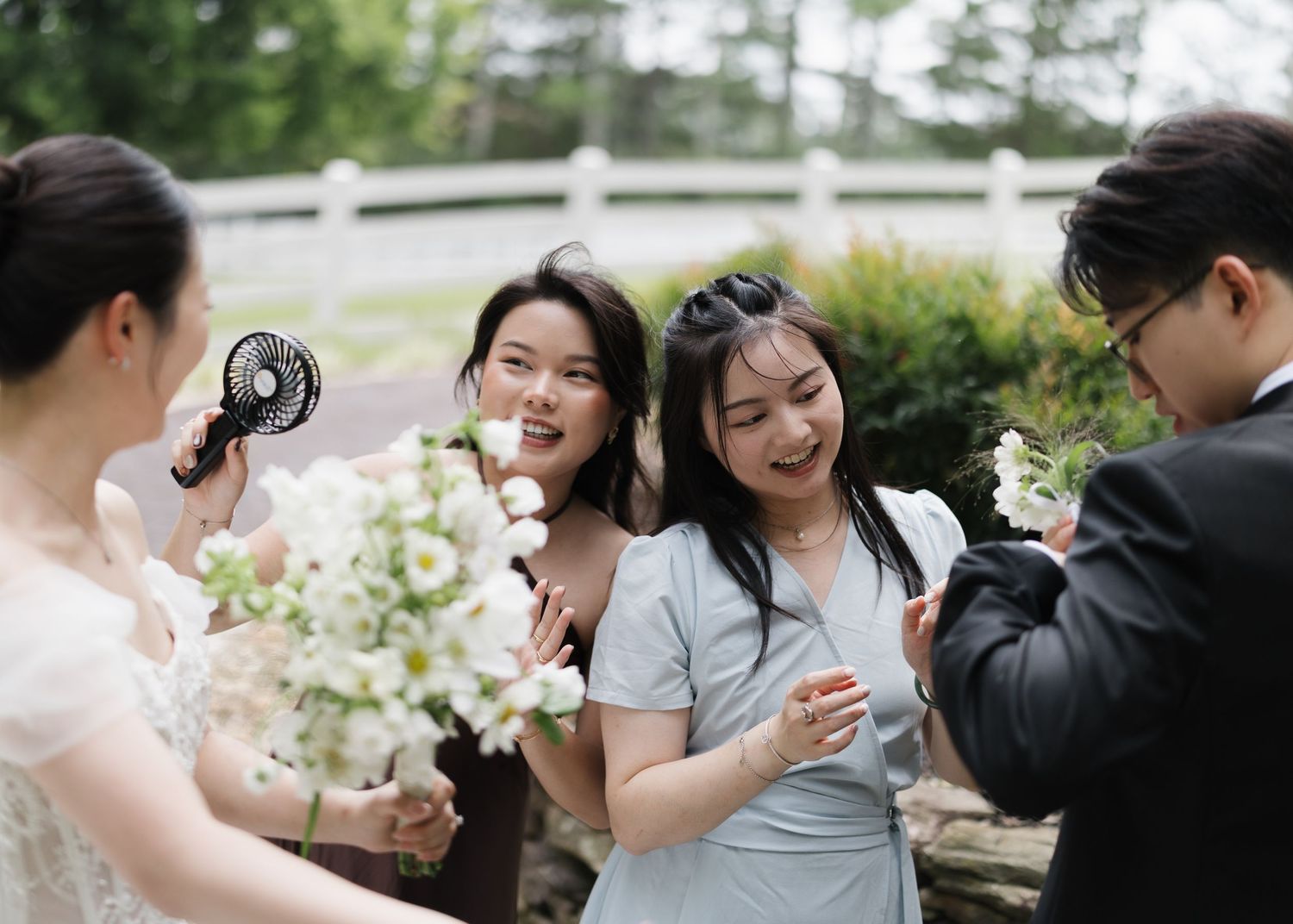 Bridesmaids laughing and using handheld fans outdoors at a wedding celebration.