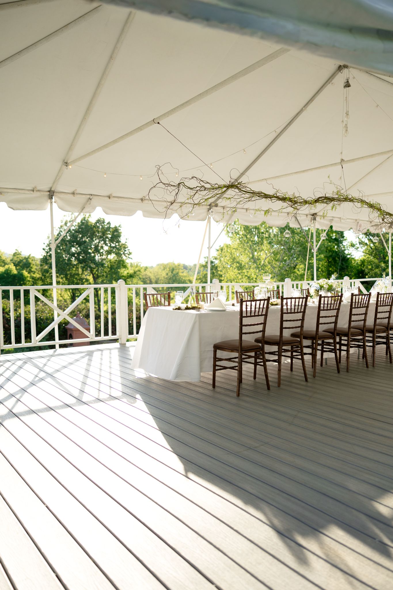 Elegant outdoor rehearsal dinner setup under white tent on wooden deck with chiavari chairs.