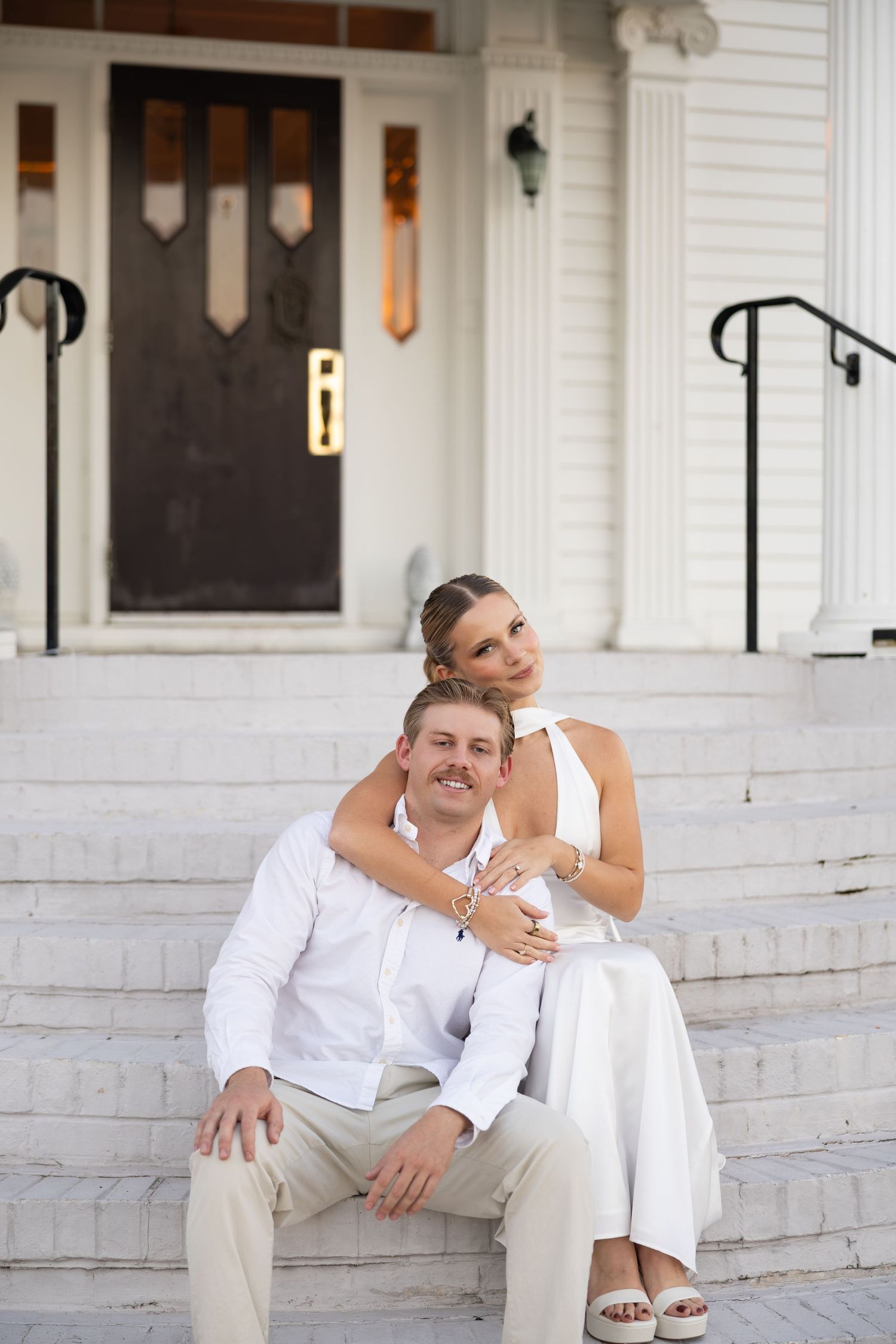 Couple in white outfits posing on steps of a white building, woman embracing man from behind.