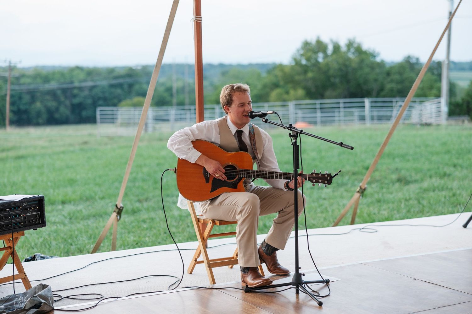 Man in suit playing acoustic guitar and singing into microphone on outdoor stage with green field backdrop.