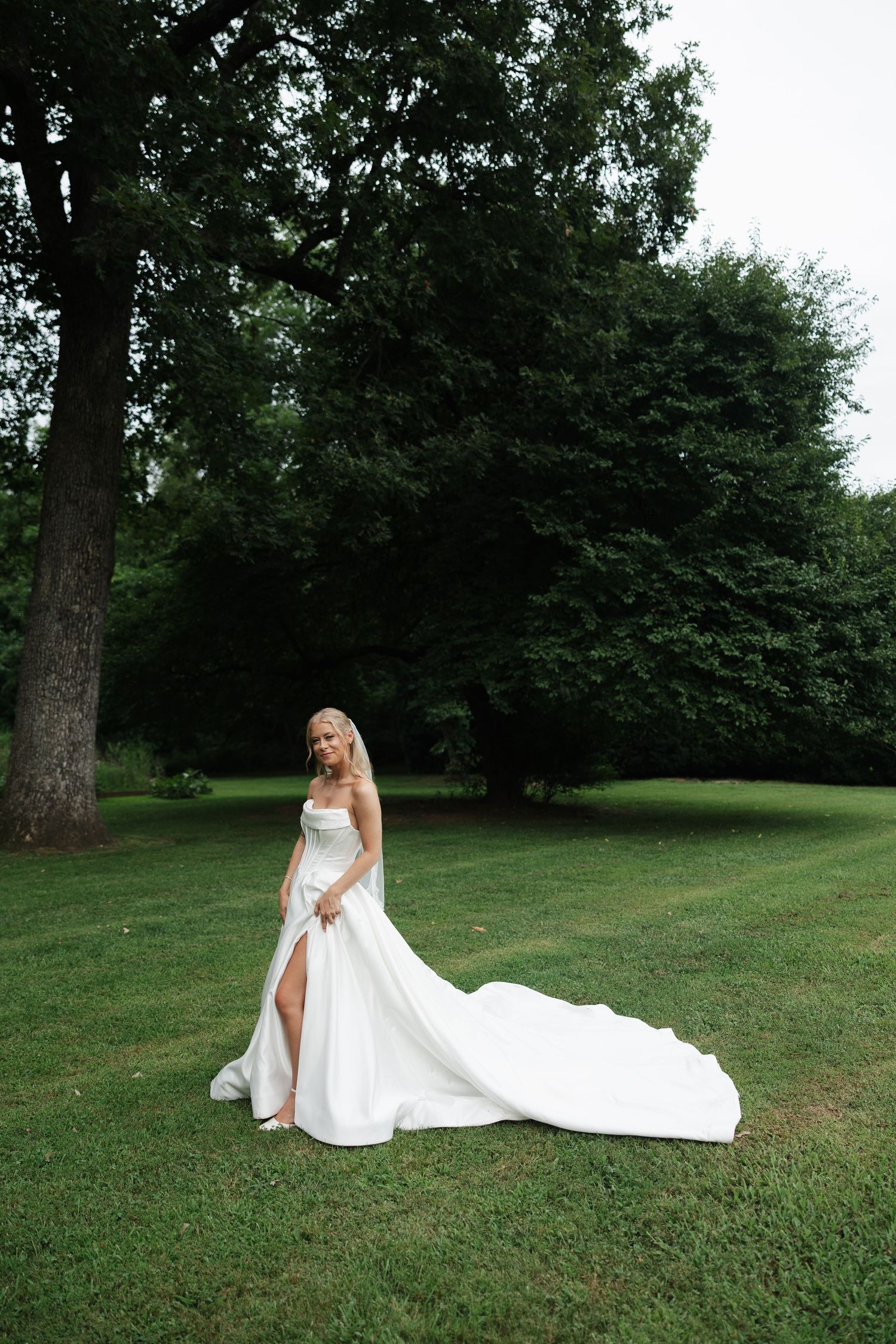 Bride in elegant white strapless gown with train poses on lush green lawn surrounded by tall trees.