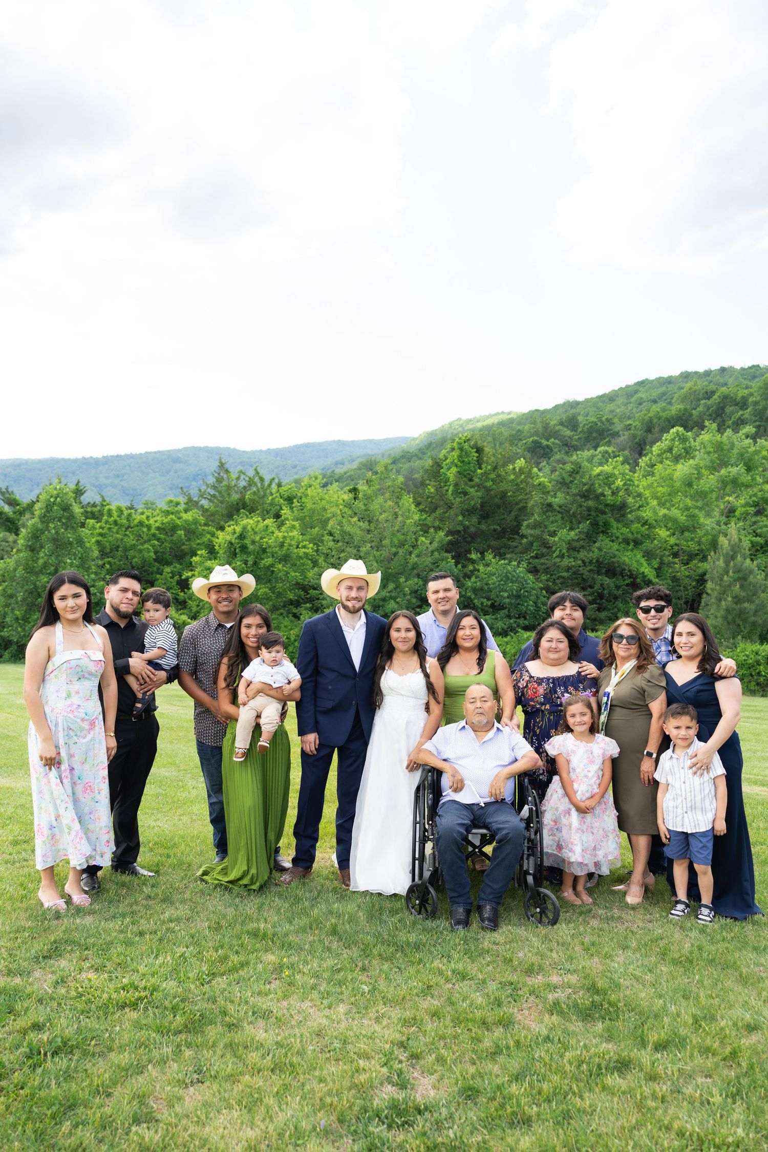 Large family group portrait at outdoor wedding with mountains in background, including bride, groom in cowboy hat, and wheelchair user.