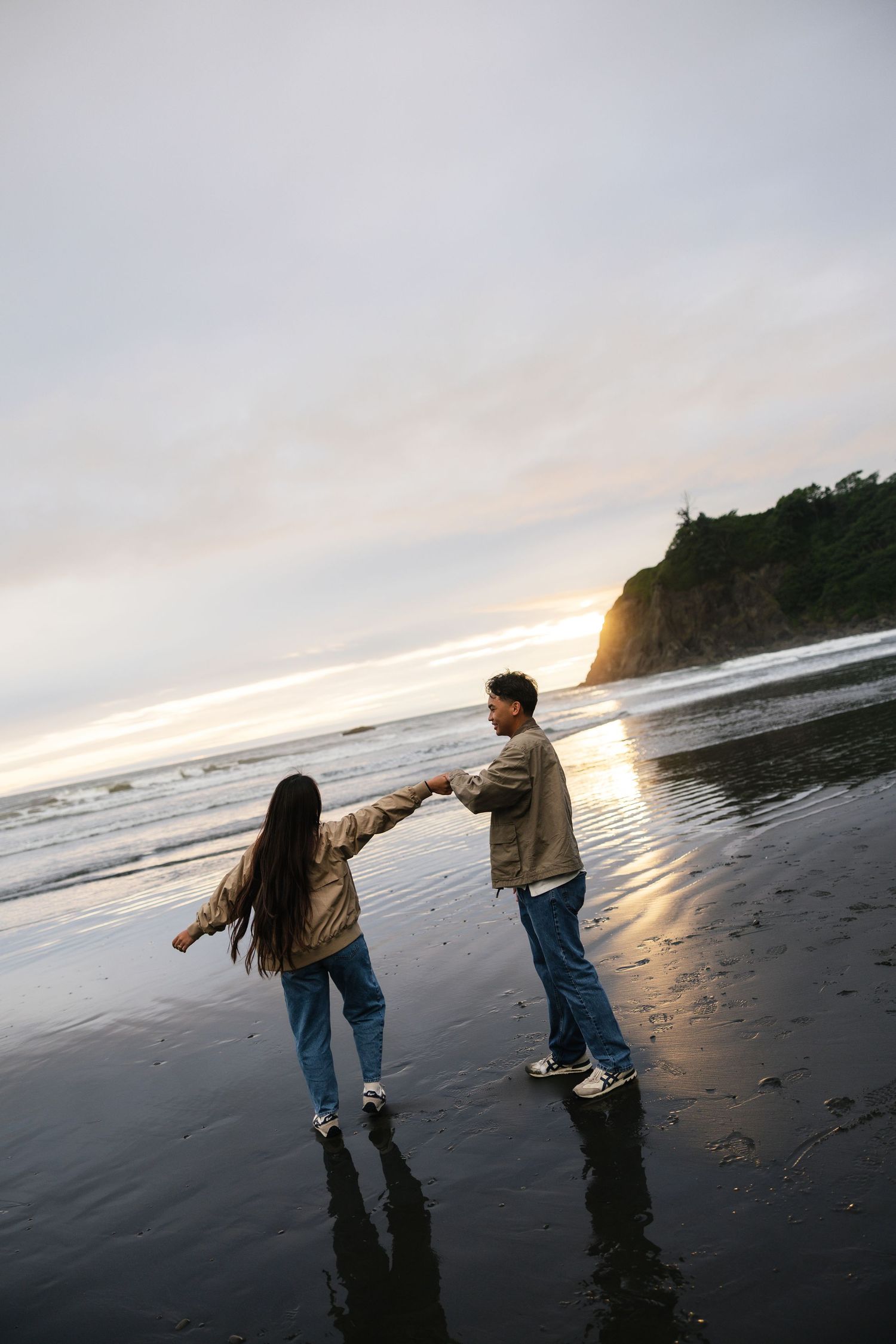 Couple holding hands on a reflective black sand beach at sunset with rocky cliffs in the background.