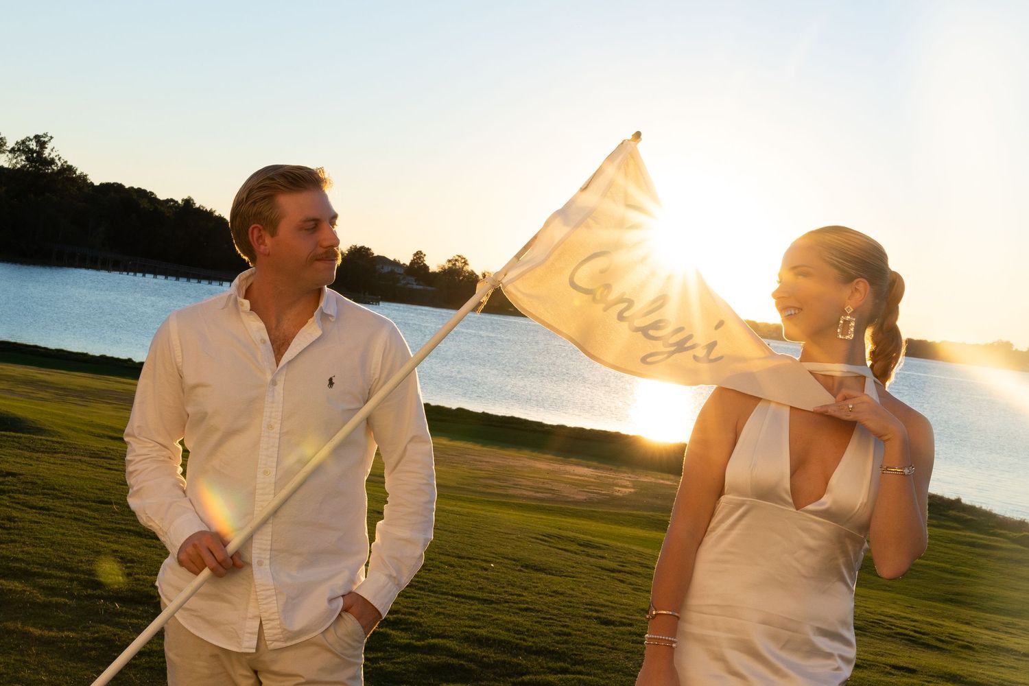 A couple walks outdoors at sunset near a lake, the bride holding a decorative white flag in warm golden light.