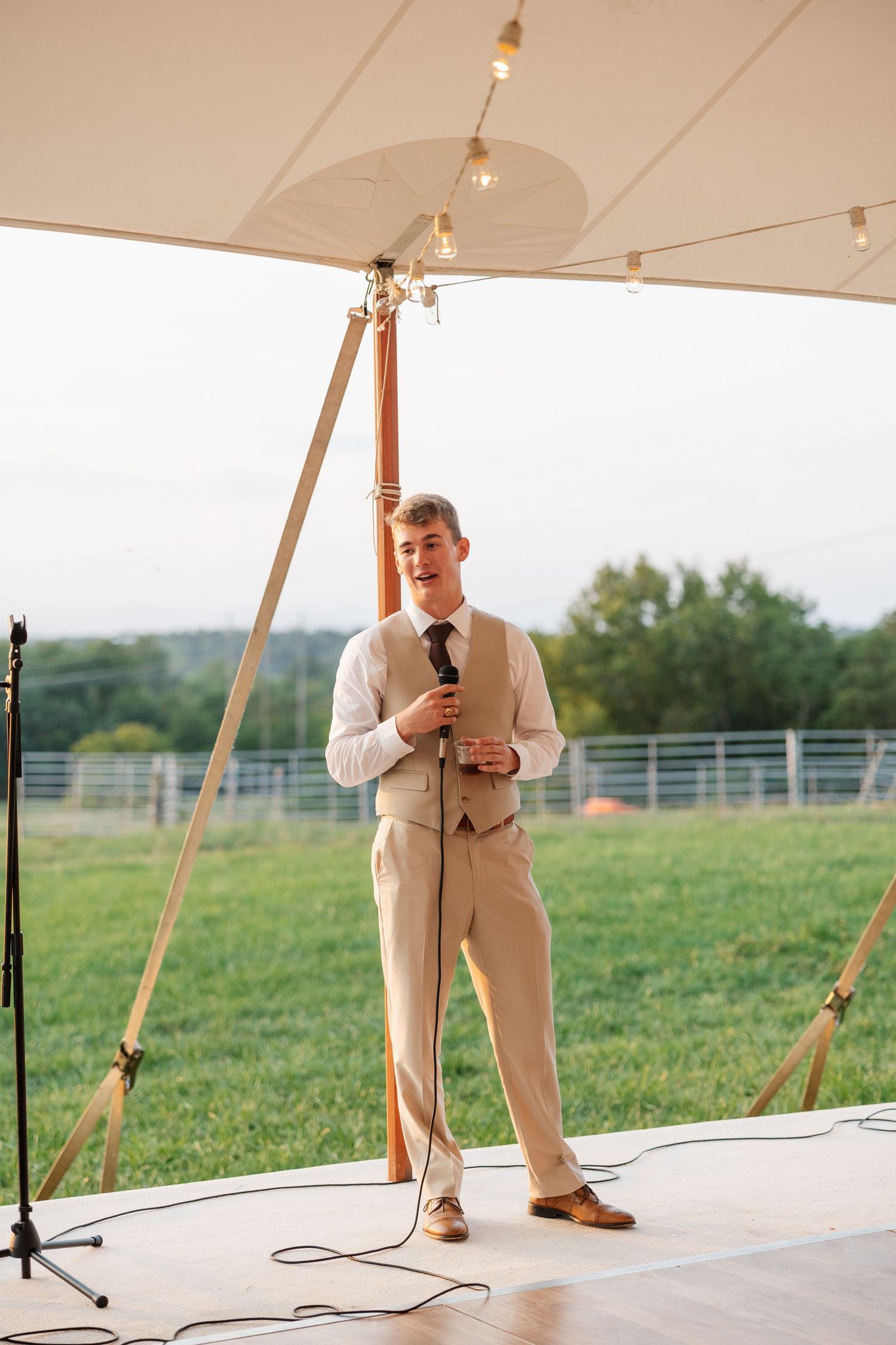 Young man in tan vest and black tie gives a speech at an outdoor wedding reception under a white tent.
