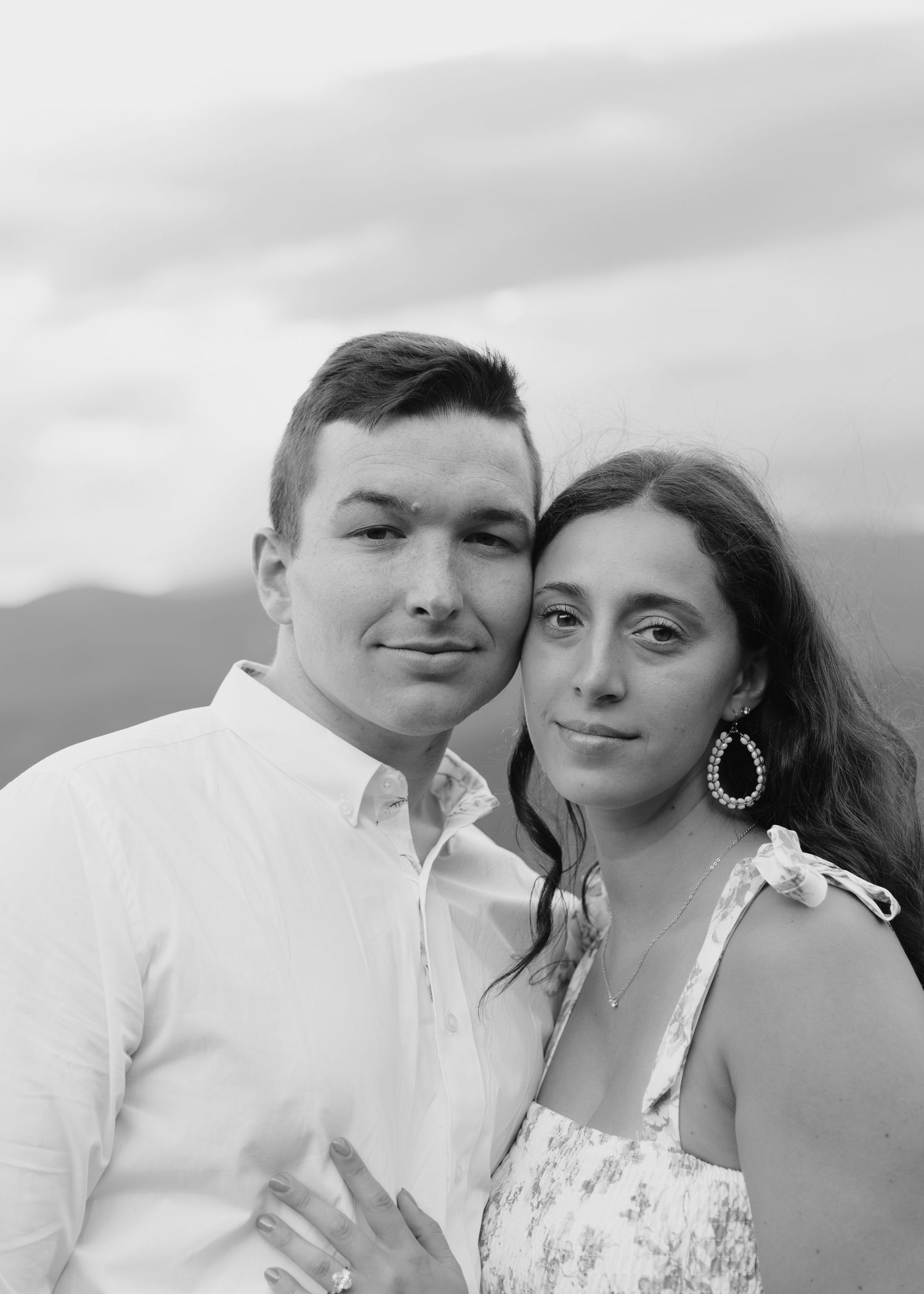 A young couple poses together in a black and white portrait with a cloudy sky and mountains behind them.