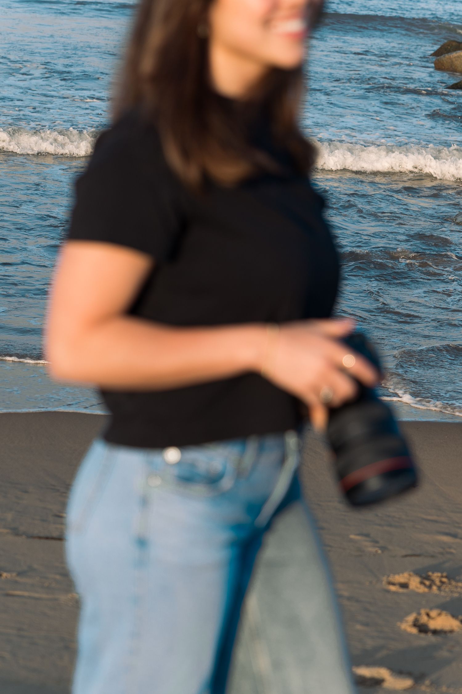 Blurred motion shot of a smiling woman in black top and jeans holding a camera walking along the beach.