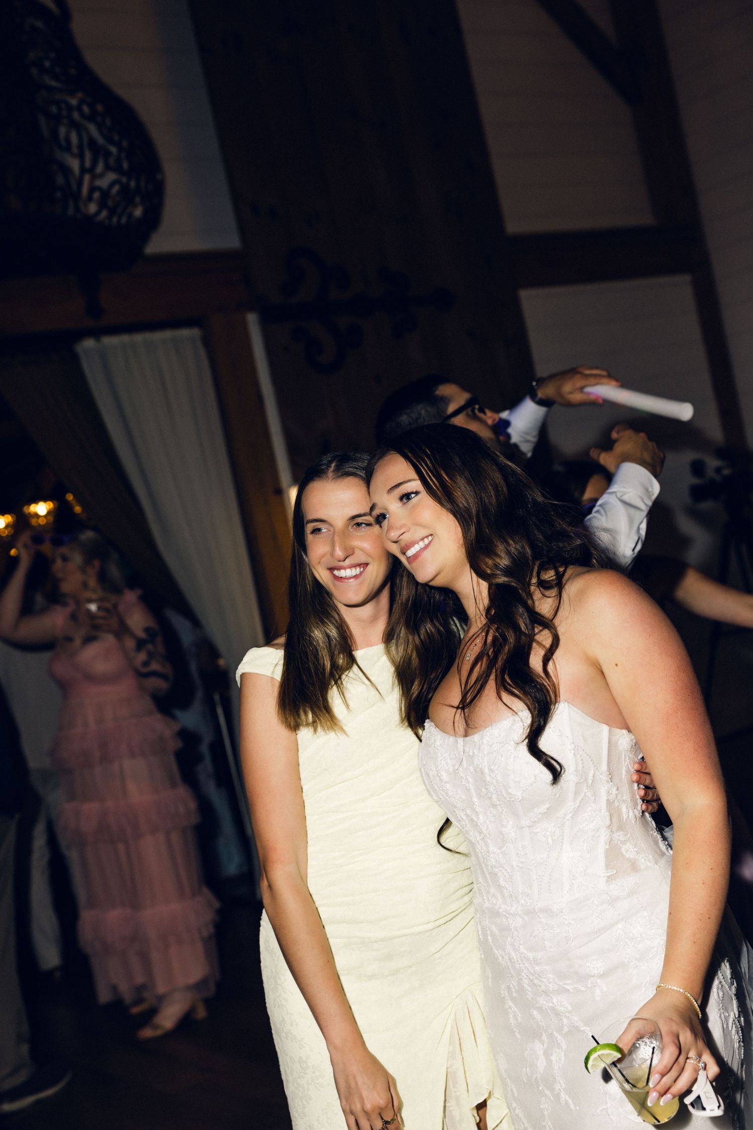 Two women in white dresses smile together at a wedding reception party.