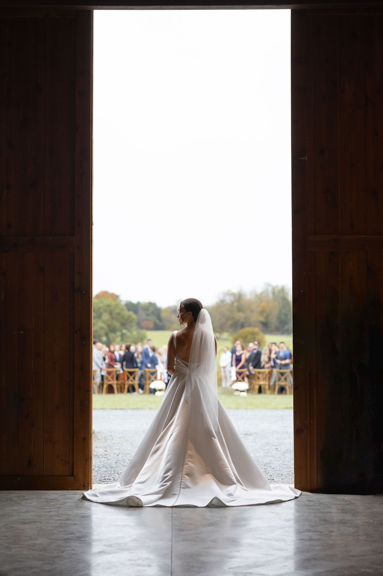 Bride in flowing white gown and veil silhouetted in large barn doors before outdoor ceremony.