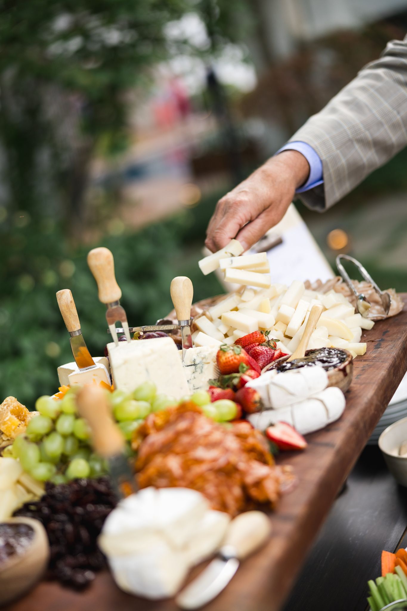 A hand reaching for cheese on an elegant charcuterie board with grapes, strawberries, and assorted cheeses at an outdoor event.