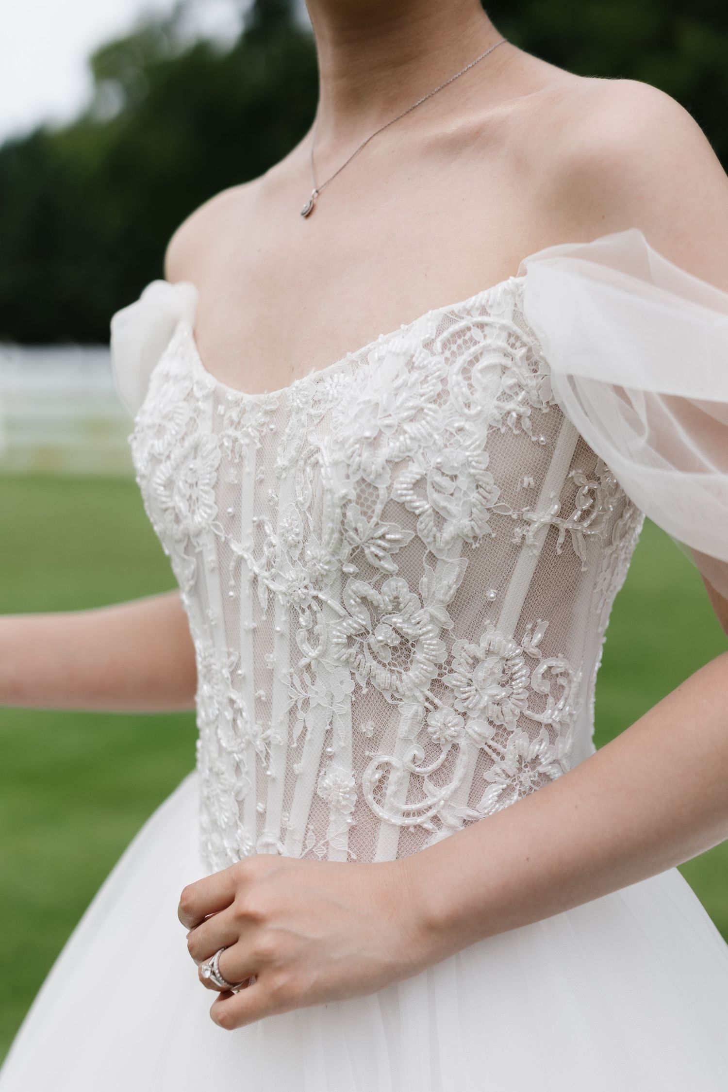 Close-up of bride's off-shoulder wedding gown featuring intricate lace applique and pearl embellishments on fitted bodice.