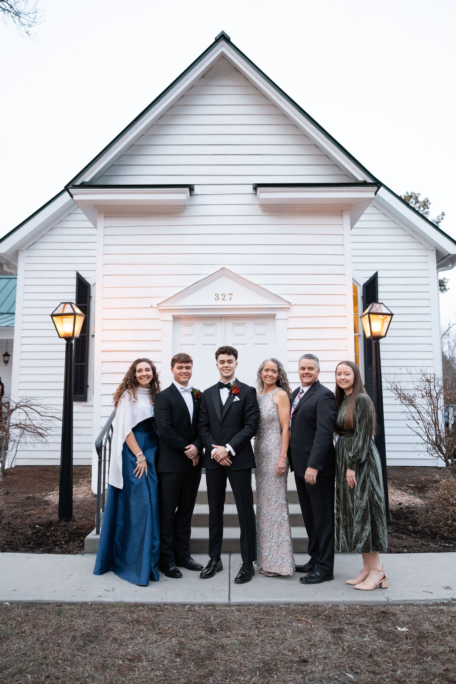 Family of six posing on steps of a white chapel with lanterns at twilight.