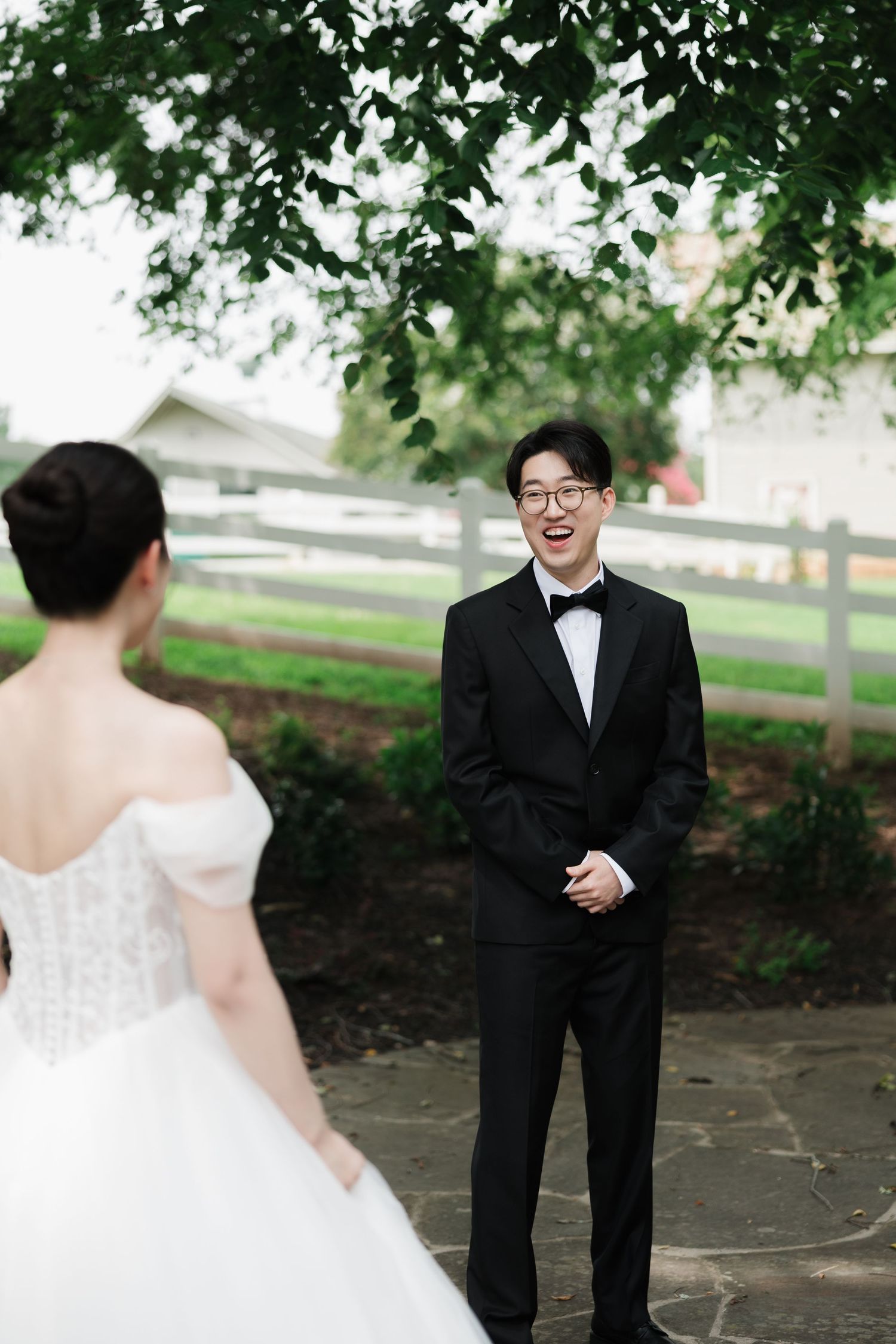 Groom in black tuxedo smiling joyfully at bride in white off-shoulder gown during outdoor first look at wedding.