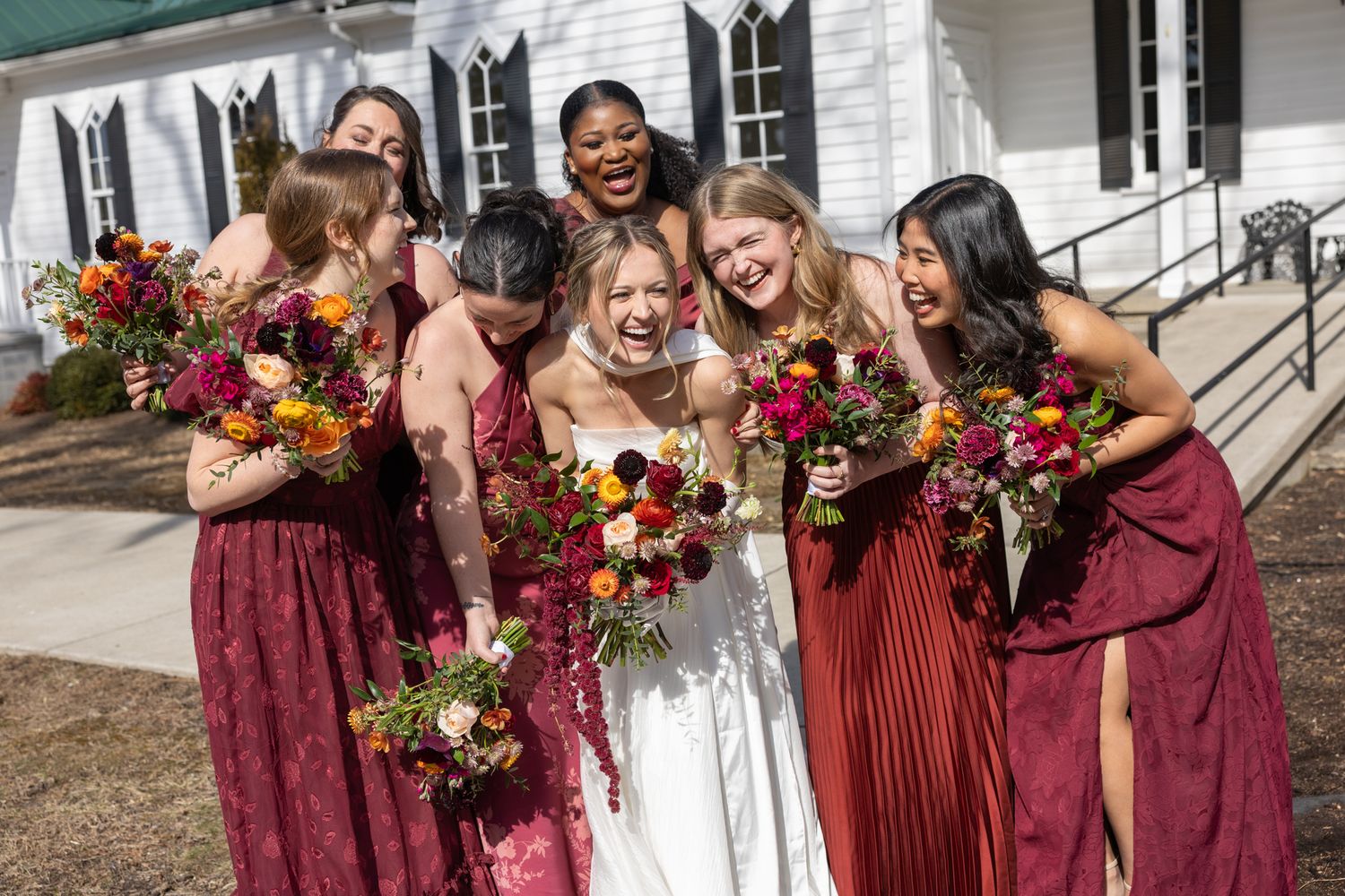 Bride laughing with bridesmaids in burgundy dresses holding vibrant bouquets outside white chapel.