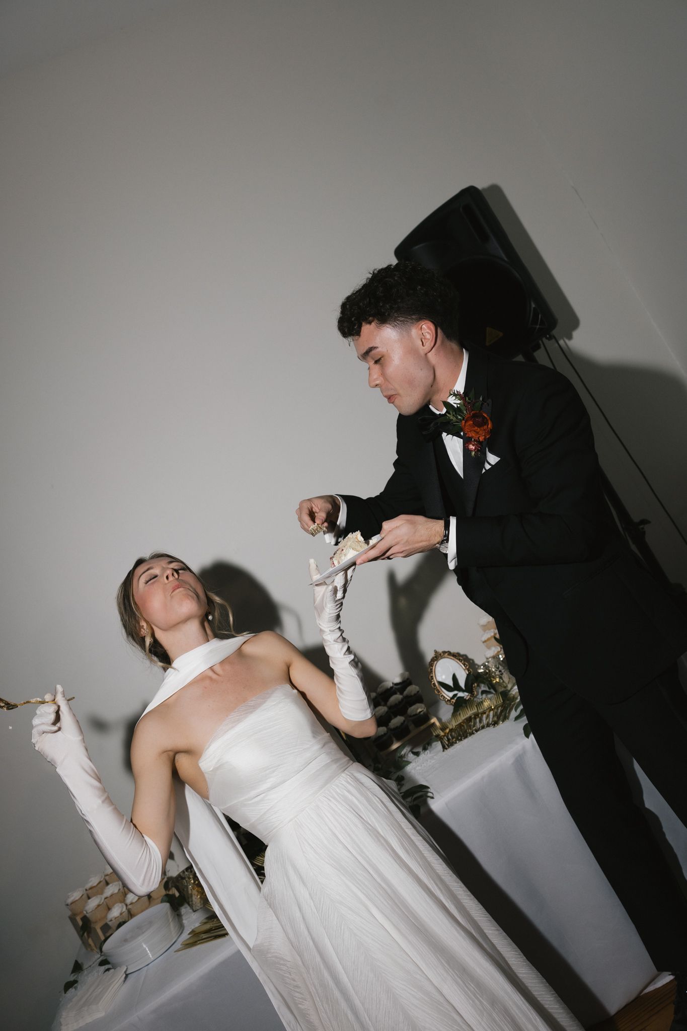 Groom in black tuxedo feeding wedding cake to bride in white gown with long gloves.
