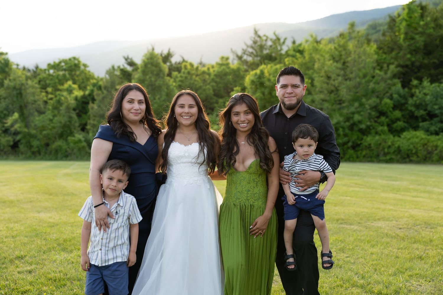 Family of six poses outdoors on green lawn with mountain backdrop, featuring woman in white dress and girl in green gown.