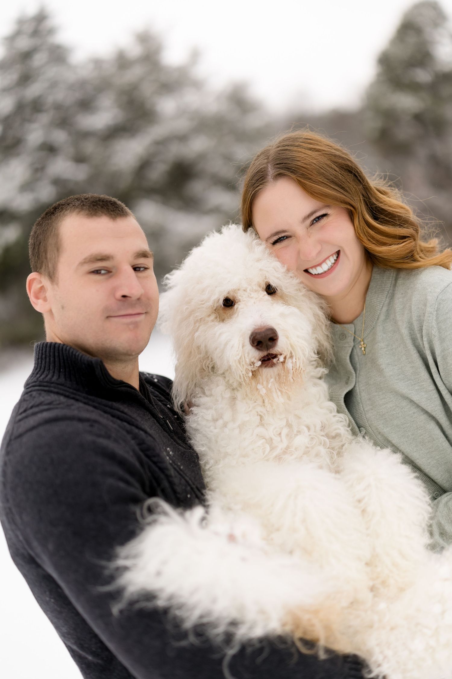 Smiling couple holding a fluffy white goldendoodle dog in a snowy outdoor winter setting.