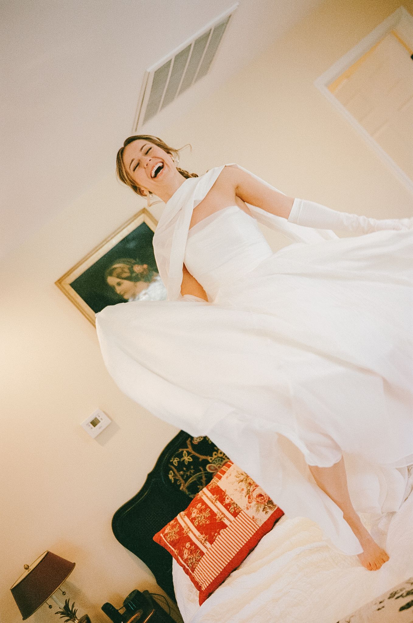 Joyful bride in white gown jumping on bed wearing long white gloves photographed on 35mm film.