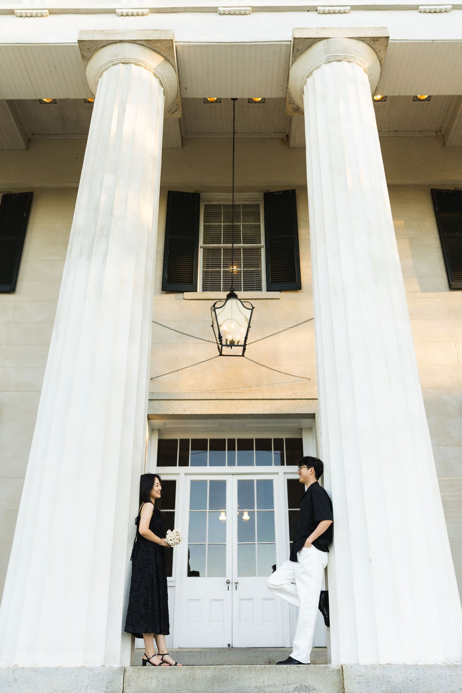 A couple in black and white outfits stands facing each other between tall white classical columns.