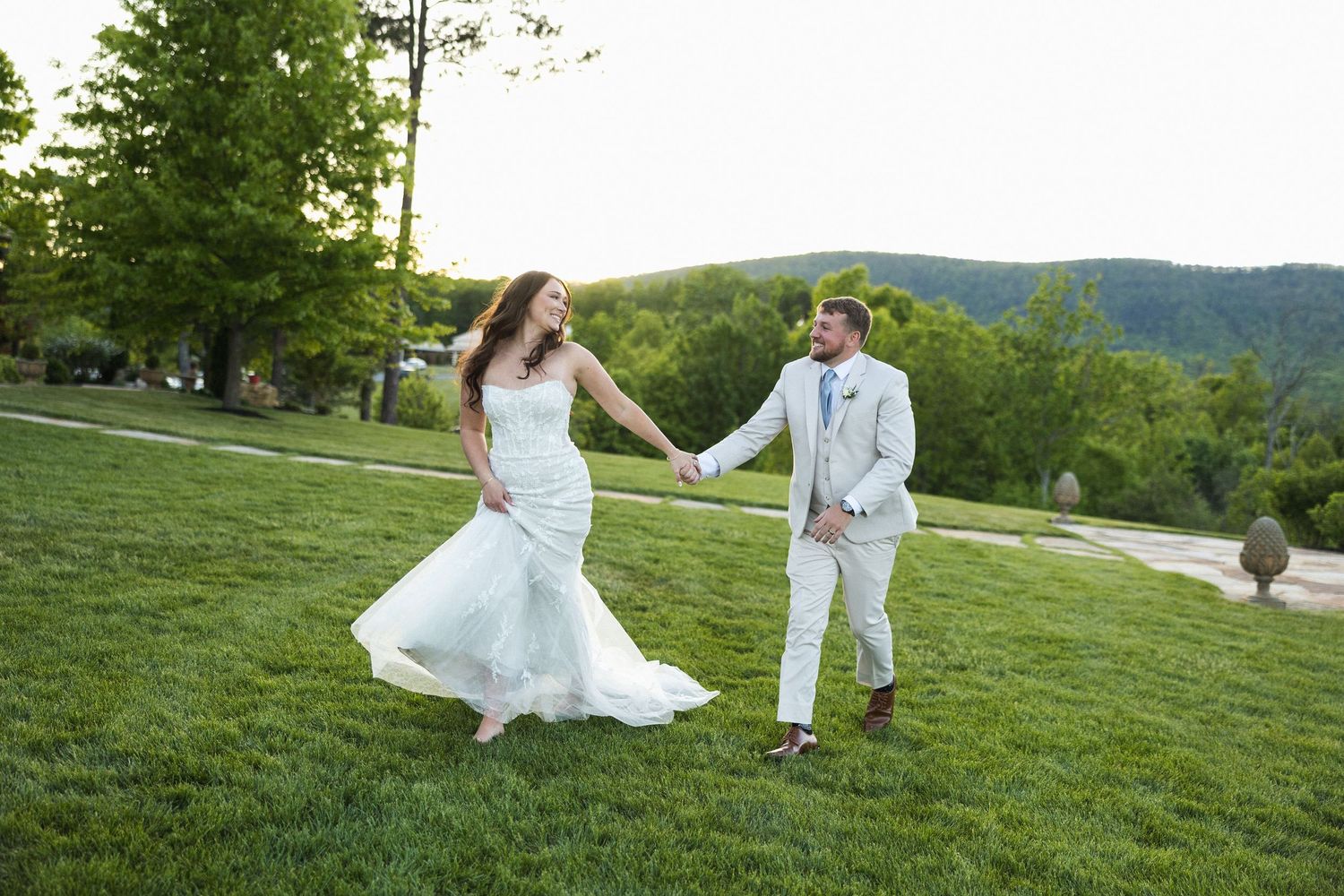Joyful bride and groom hold hands running across green lawn with mountain scenery.
