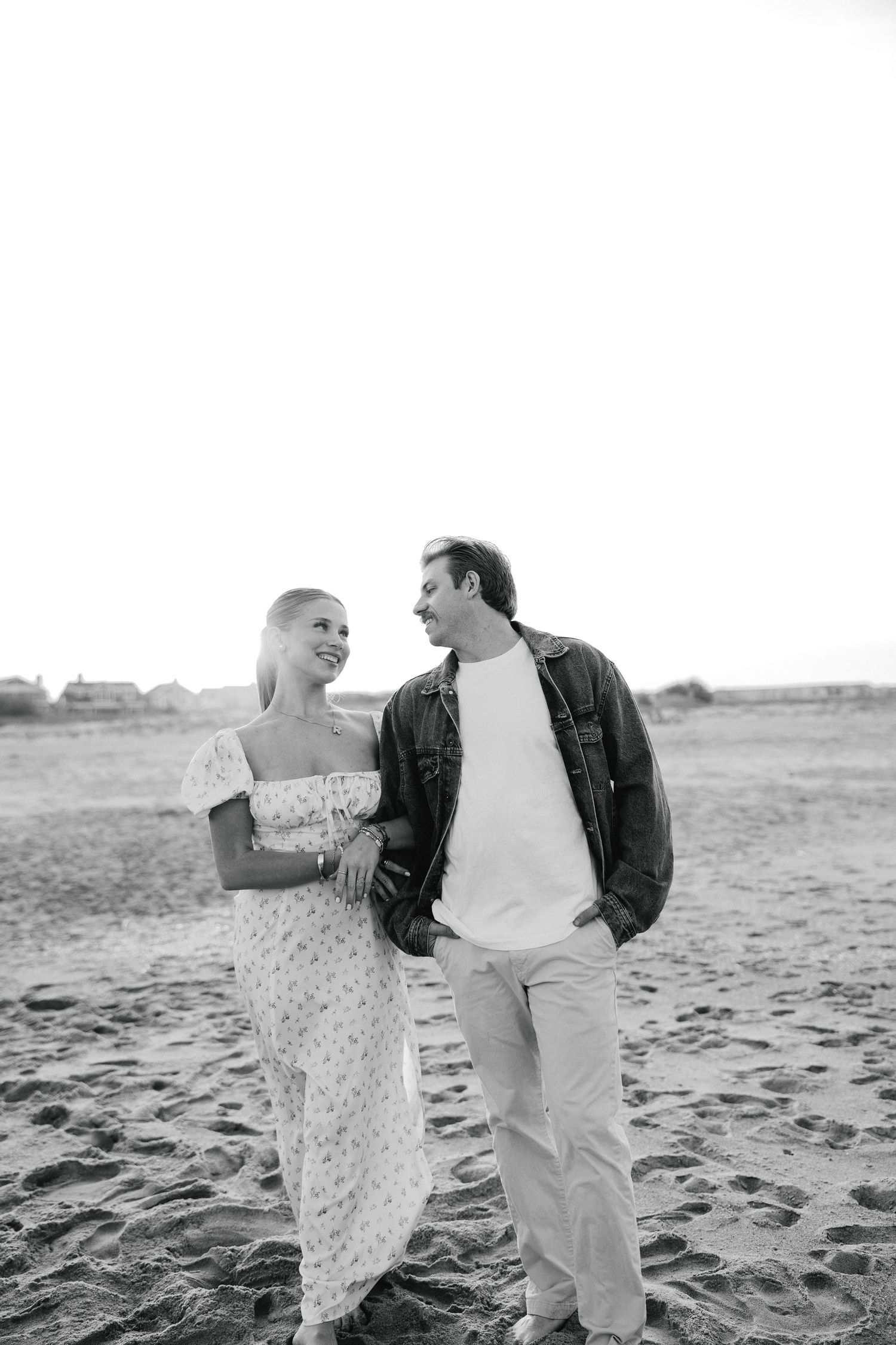 Black and white photo of smiling couple walking together on sandy beach, woman in floral dress.