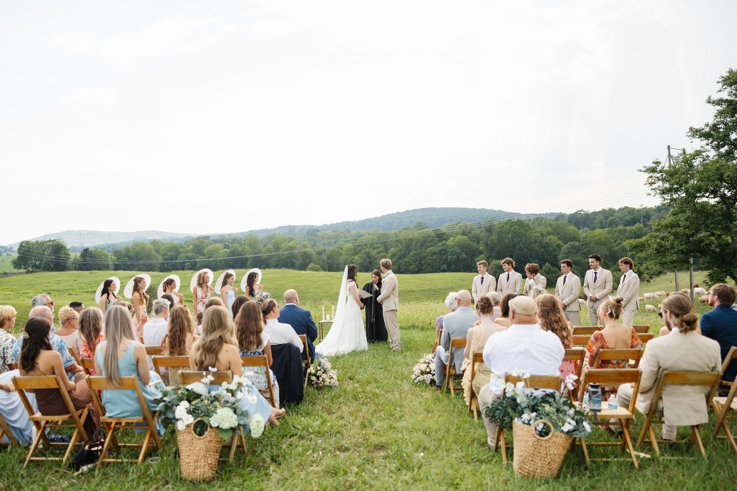 Outdoor wedding ceremony in a green meadow with mountain views, guests seated in wooden chairs with floral basket arrangements.