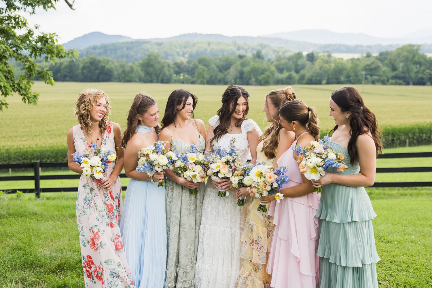 Six bridesmaids in mismatched pastel floral gowns holding bouquets of wildflowers on a lush green farm with mountain views.