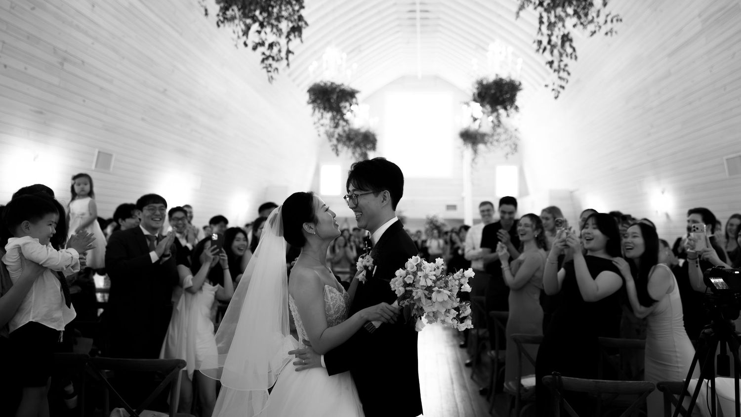 Black and white wedding ceremony photo of couple sharing first kiss at altar surrounded by cheering guests in elegant chapel.