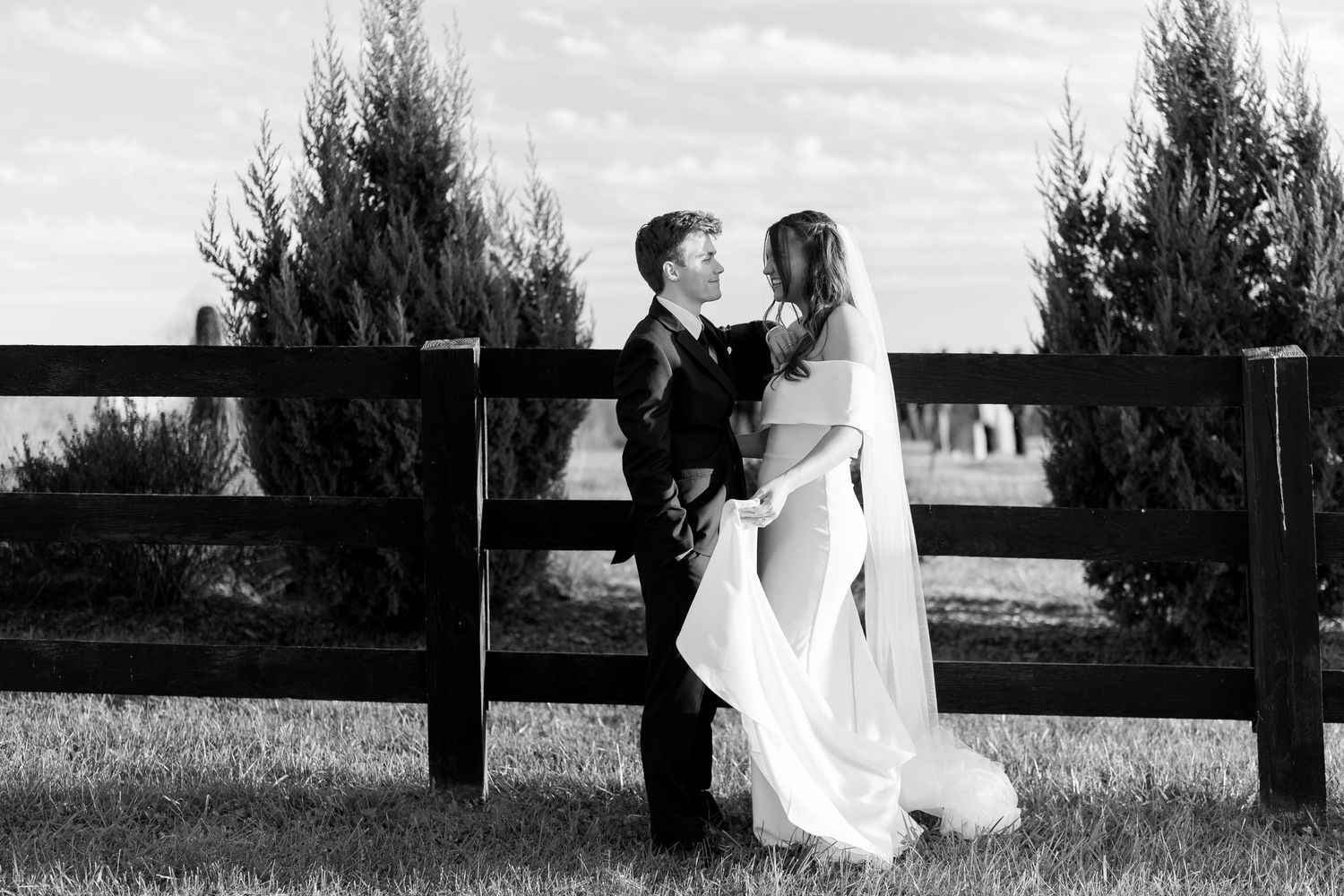 Black and white photo of bride and groom sharing a romantic moment by a wooden fence surrounded by evergreen trees.