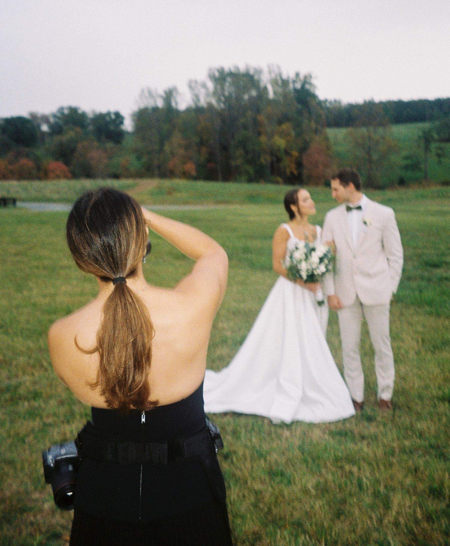 Photographer capturing bride and groom posing in a green meadow on their wedding day.