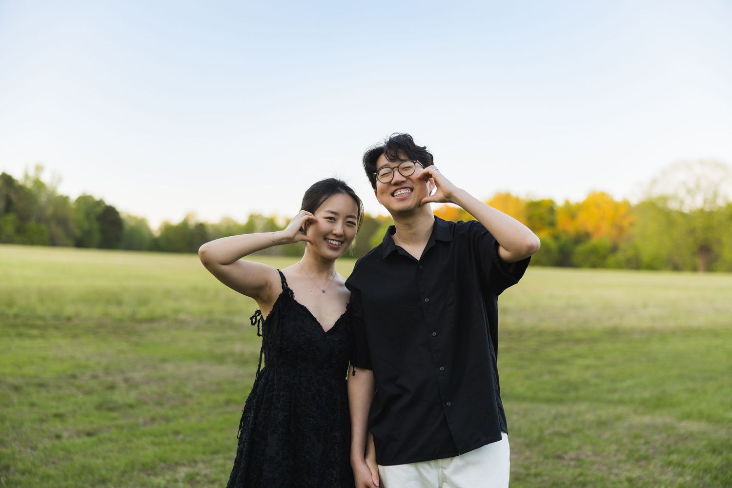 A cheerful couple makes peace signs and smiles in an open green field during a sunny outdoor photo session.