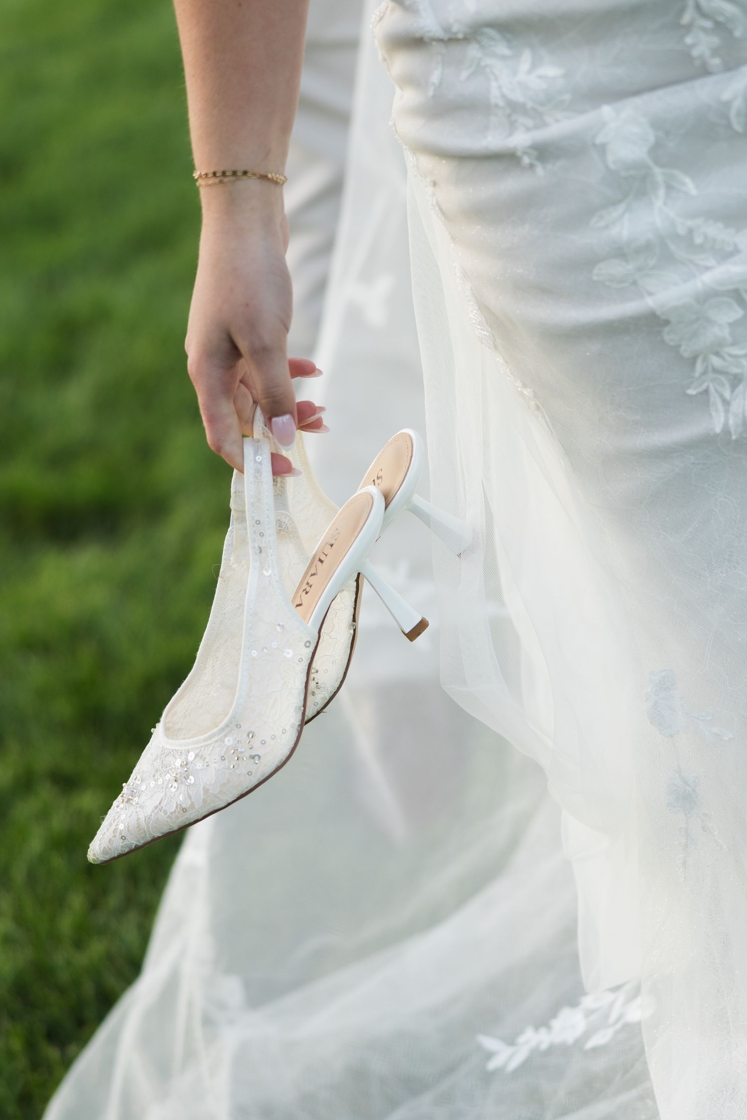 Bride in white lace wedding gown holds elegant embellished pointed-toe bridal heel shoe on green grass lawn.
