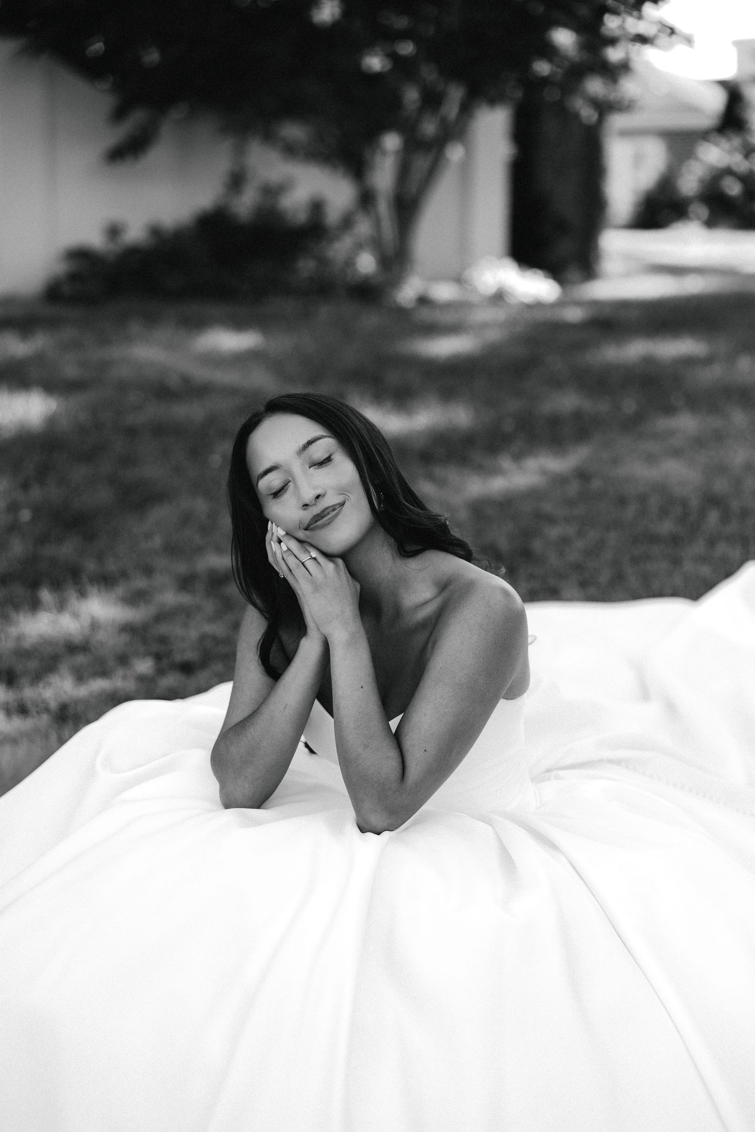 Black and white photo of a bride in a white gown lying on grass, hands folded near her face, smiling softly.