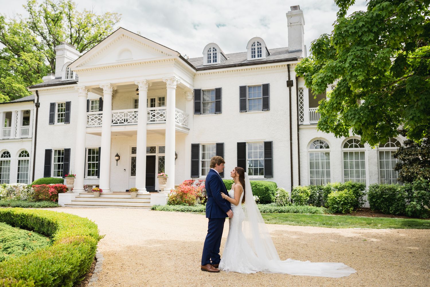Bride and groom share a romantic moment outside a grand white colonial mansion with black shutters and lush gardens.
