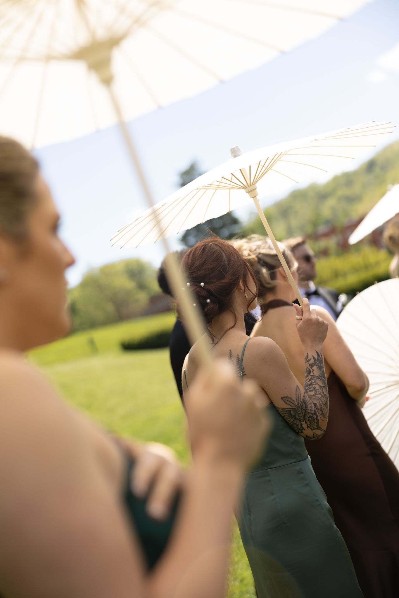 Wedding guests holding white parasols outdoors on a sunny day, bridesmaid with floral tattoo visible.