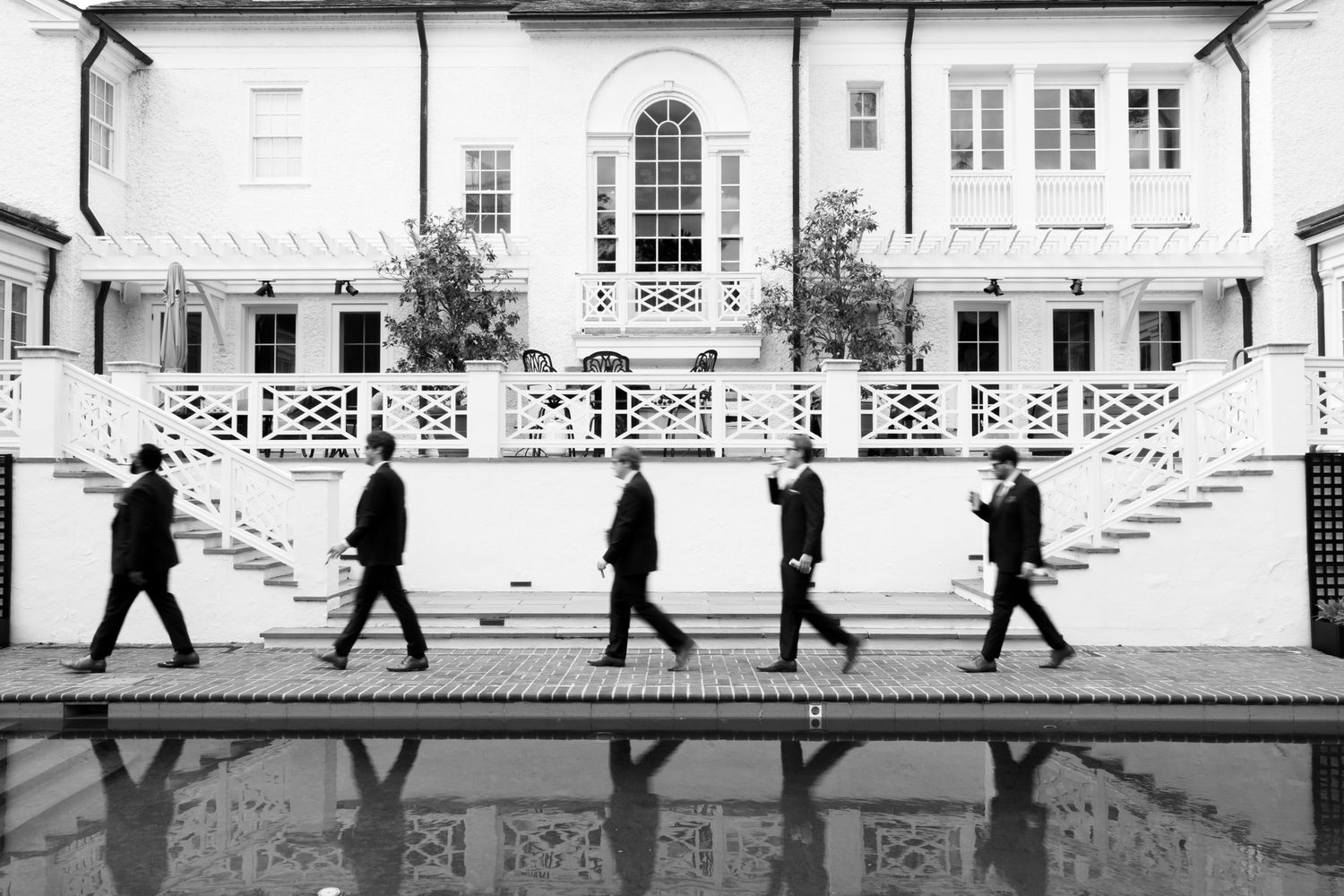 Groomsmen in black suits walk in a line beside a pool reflecting a white mansion in black and white photo.