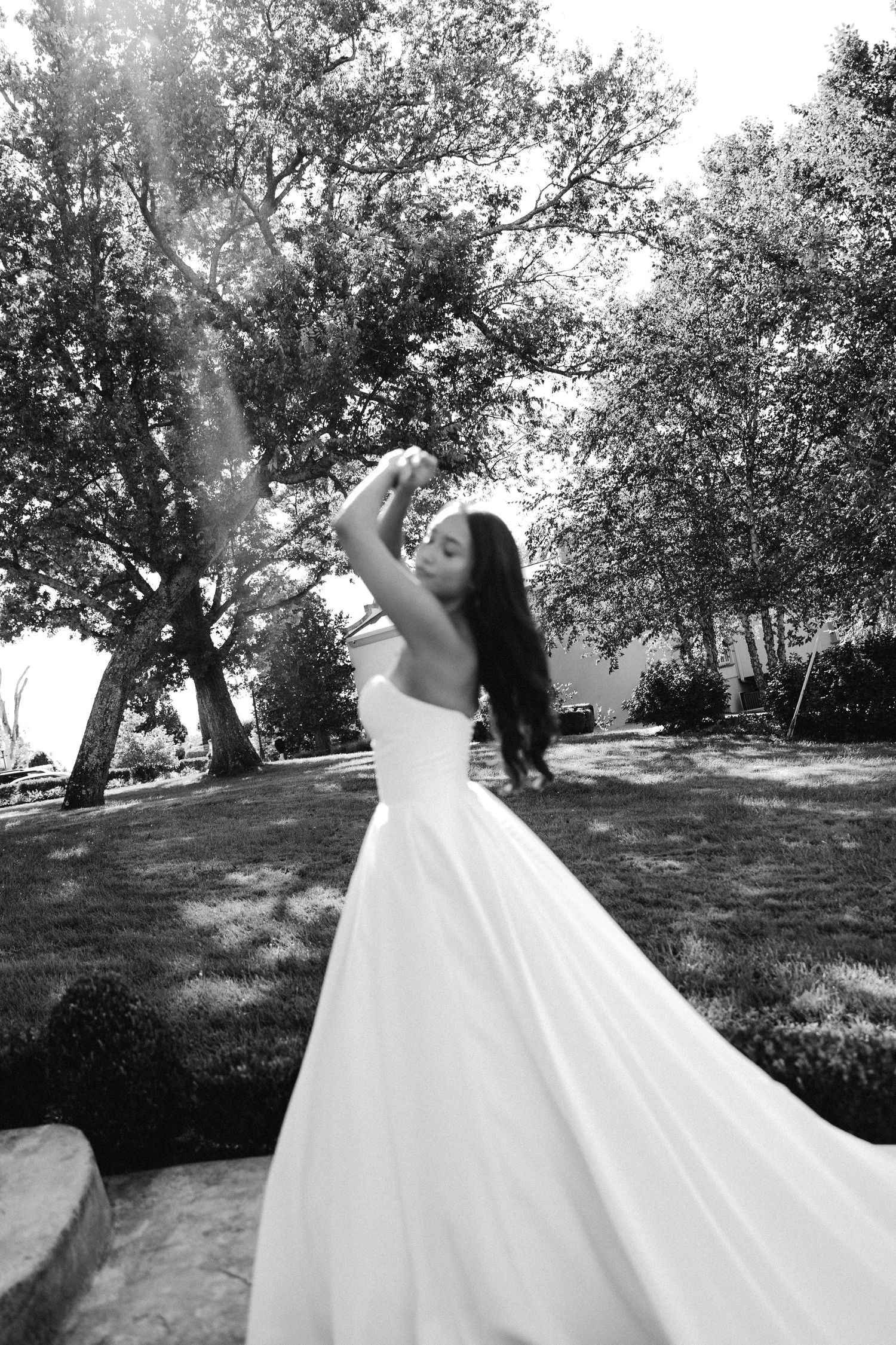 Black and white photo of a bride in a flowing white gown posing gracefully outdoors among lush trees.
