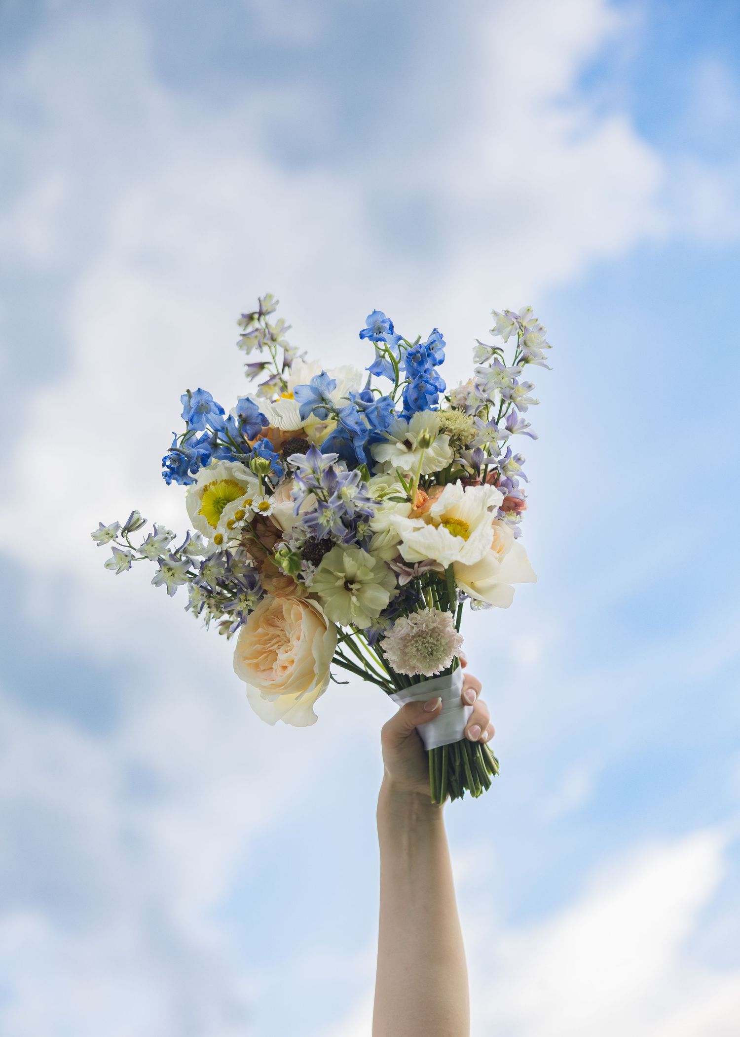 Hand holding a beautiful blue and white wedding bouquet with roses and delphiniums against a cloudy sky.