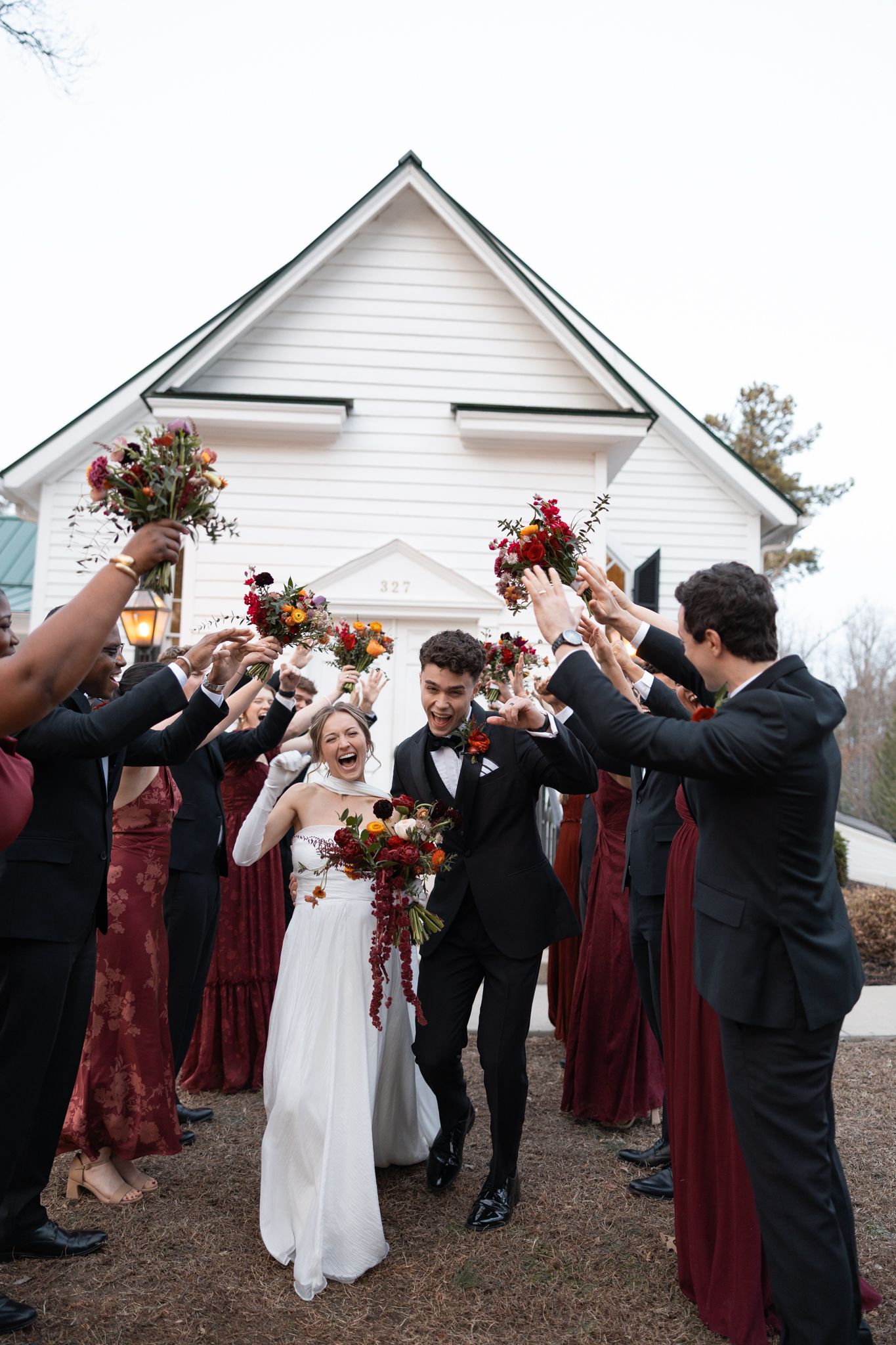 Bride and groom exit white chapel through cheering wedding party raising colorful autumn bouquets.
