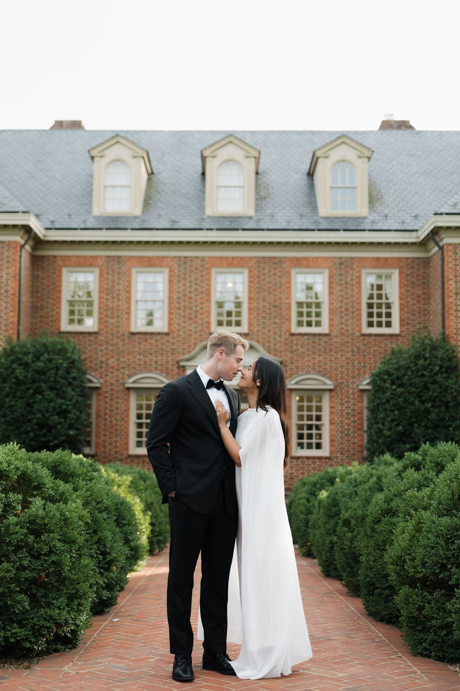 Bride and groom share a romantic kiss outside a grand brick mansion surrounded by manicured hedges.