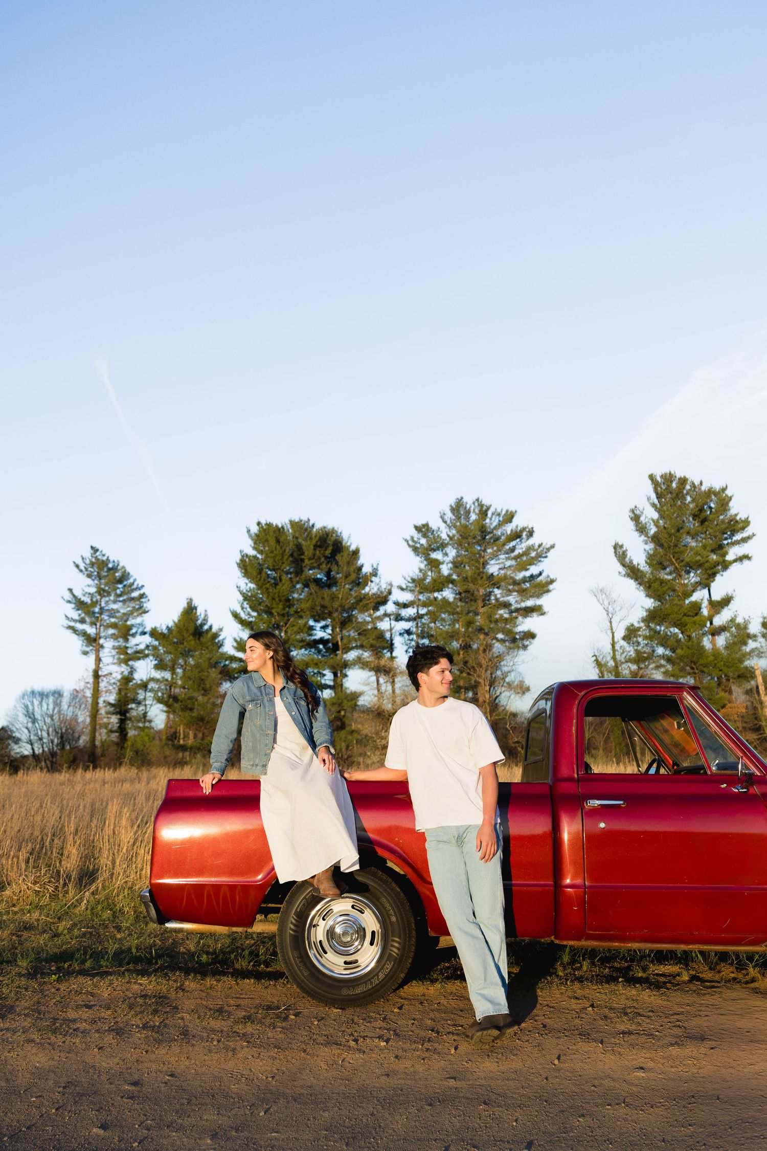 A couple standing apart beside a red vintage pickup truck on a dirt road, surrounded by tall pine trees.