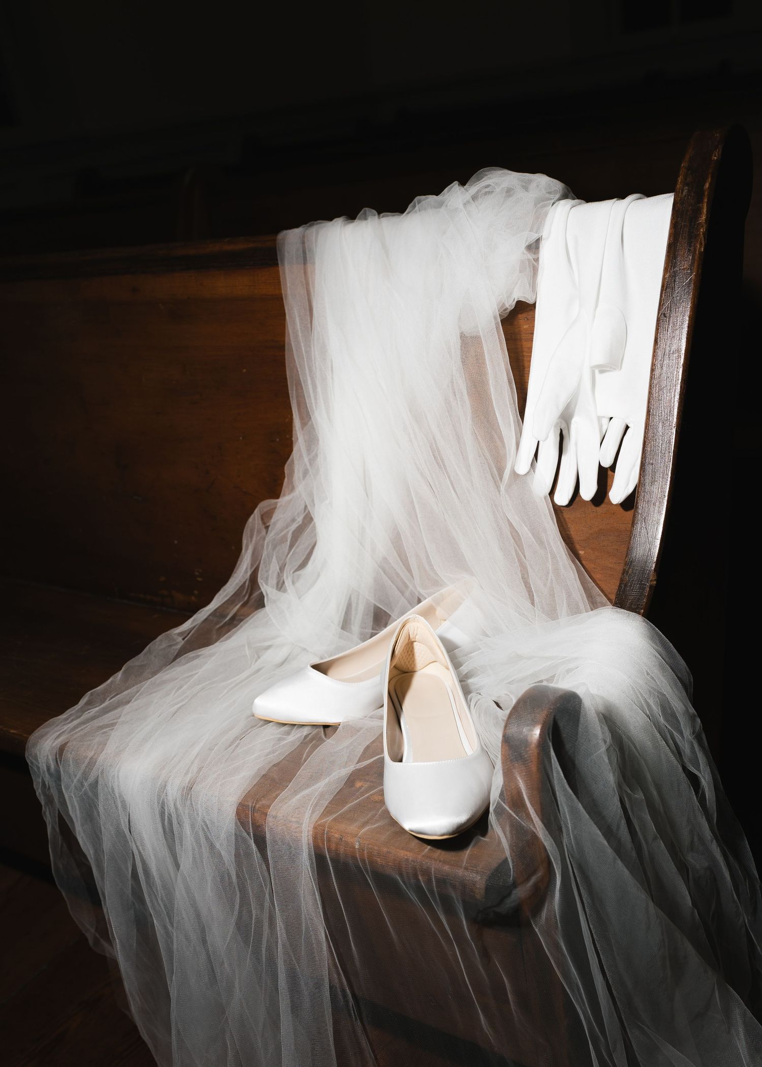 Elegant bridal accessories including white heels, lace gloves, and tulle veil on vintage wooden chair.