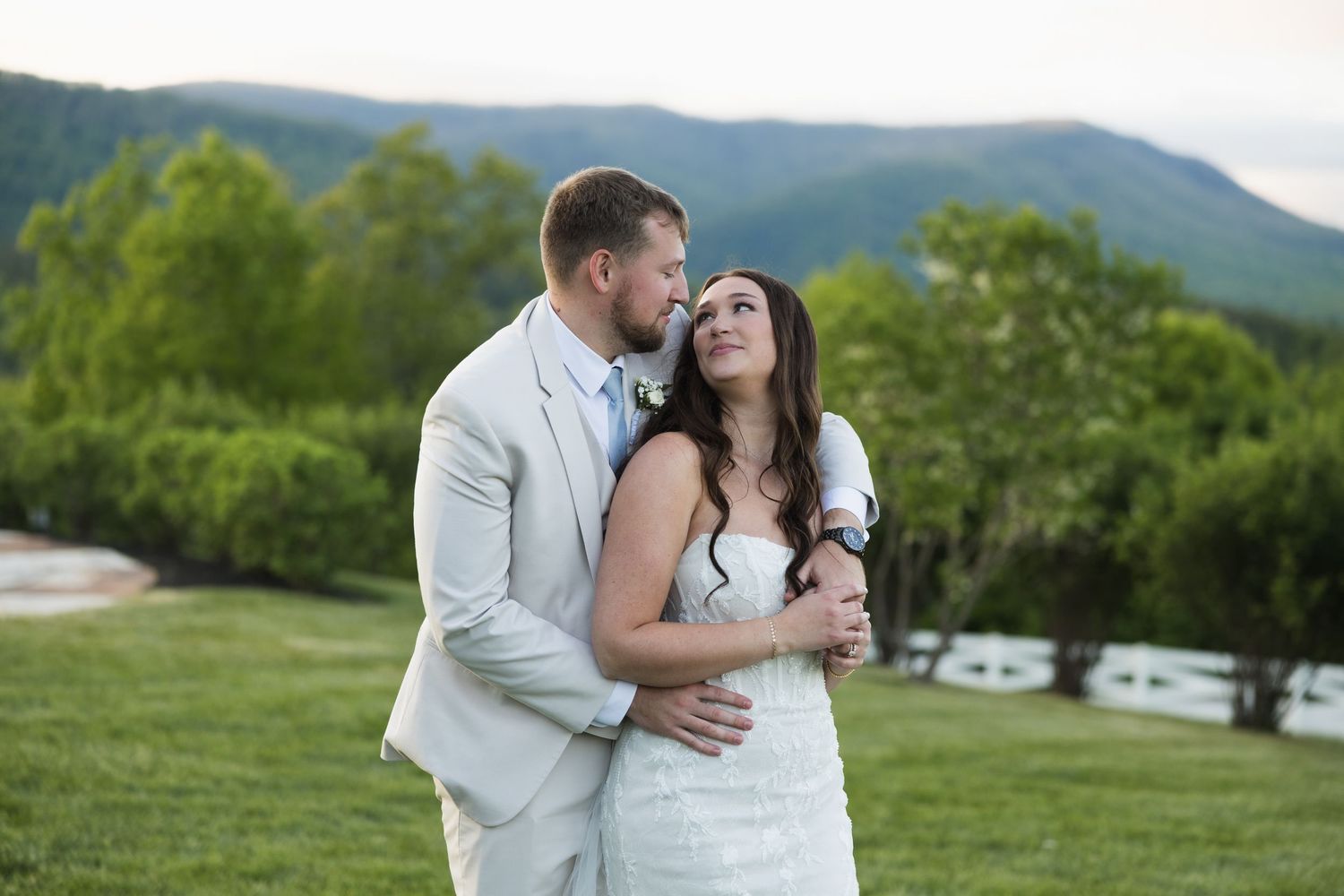 Romantic bride and groom embrace outdoors at sunset with scenic blue mountain range and green hills behind them.