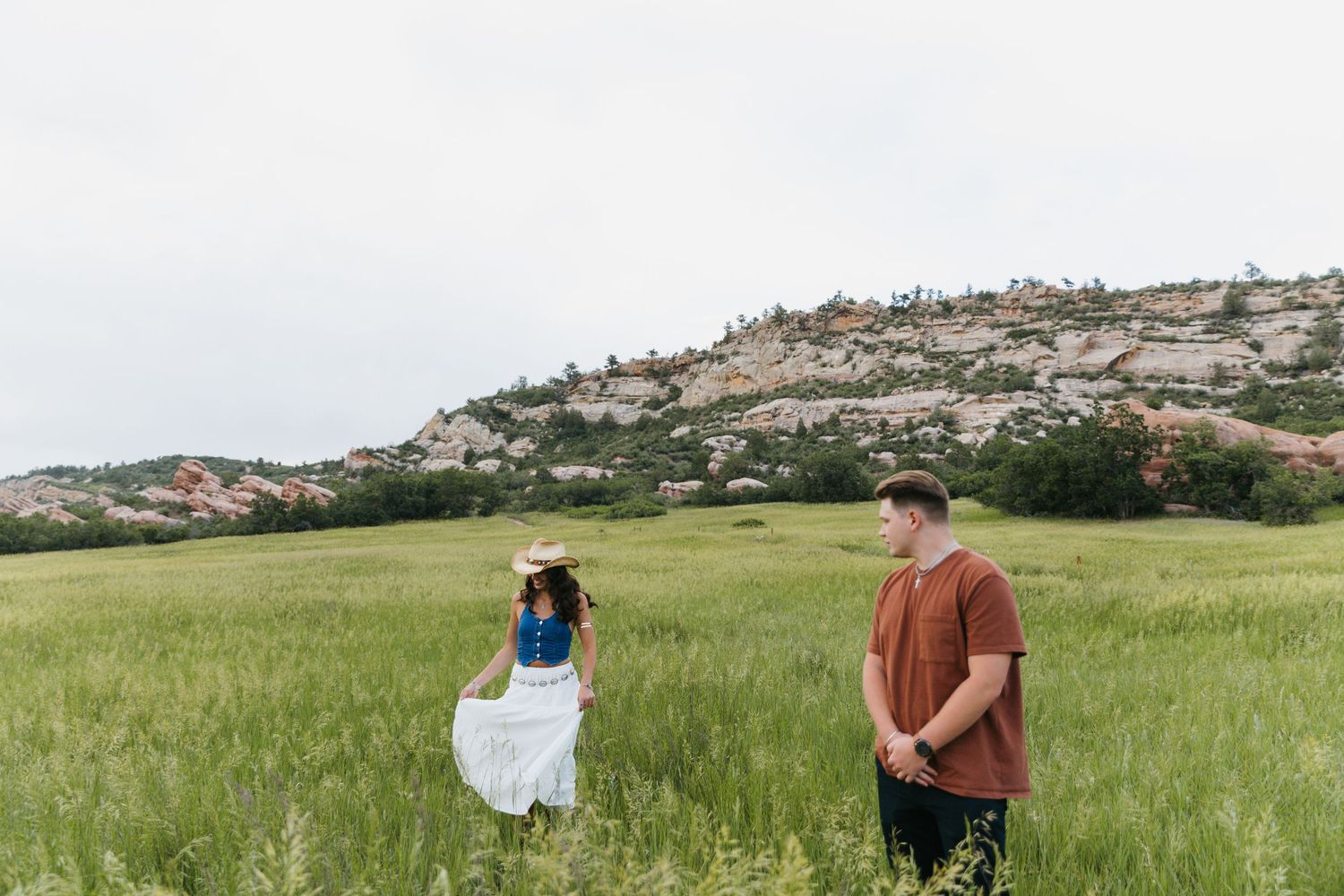 Couple standing in green meadow with rocky mountain landscape in the background.